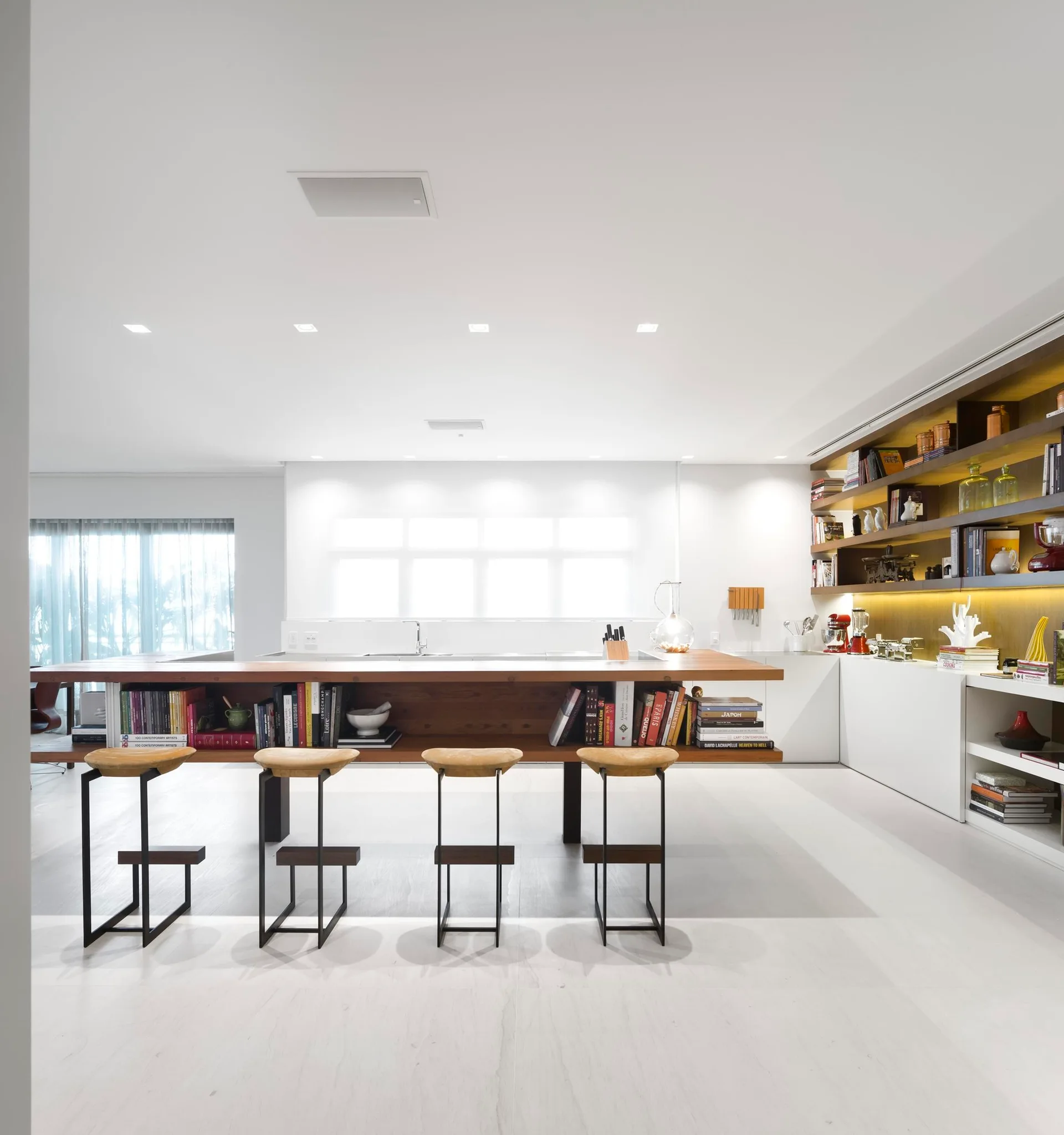 A minimalist white kitchen features a long wood island with integrated book storage, four bar stools, and warm backlit shelving.