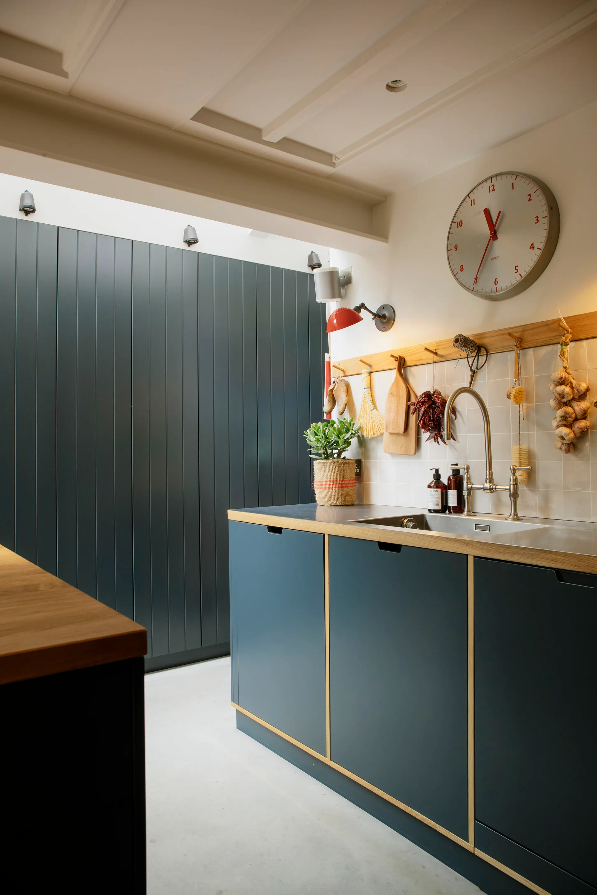 Dark blue kitchen cabinets with wood trim sit below a tiled backsplash, a stainless steel sink, and a wooden peg rail hanging kitchen tools.