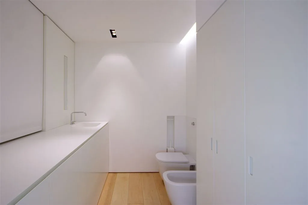 A minimalist white bathroom features a long seamless vanity, a wall-mounted toilet and bidet, and light wood plank flooring.