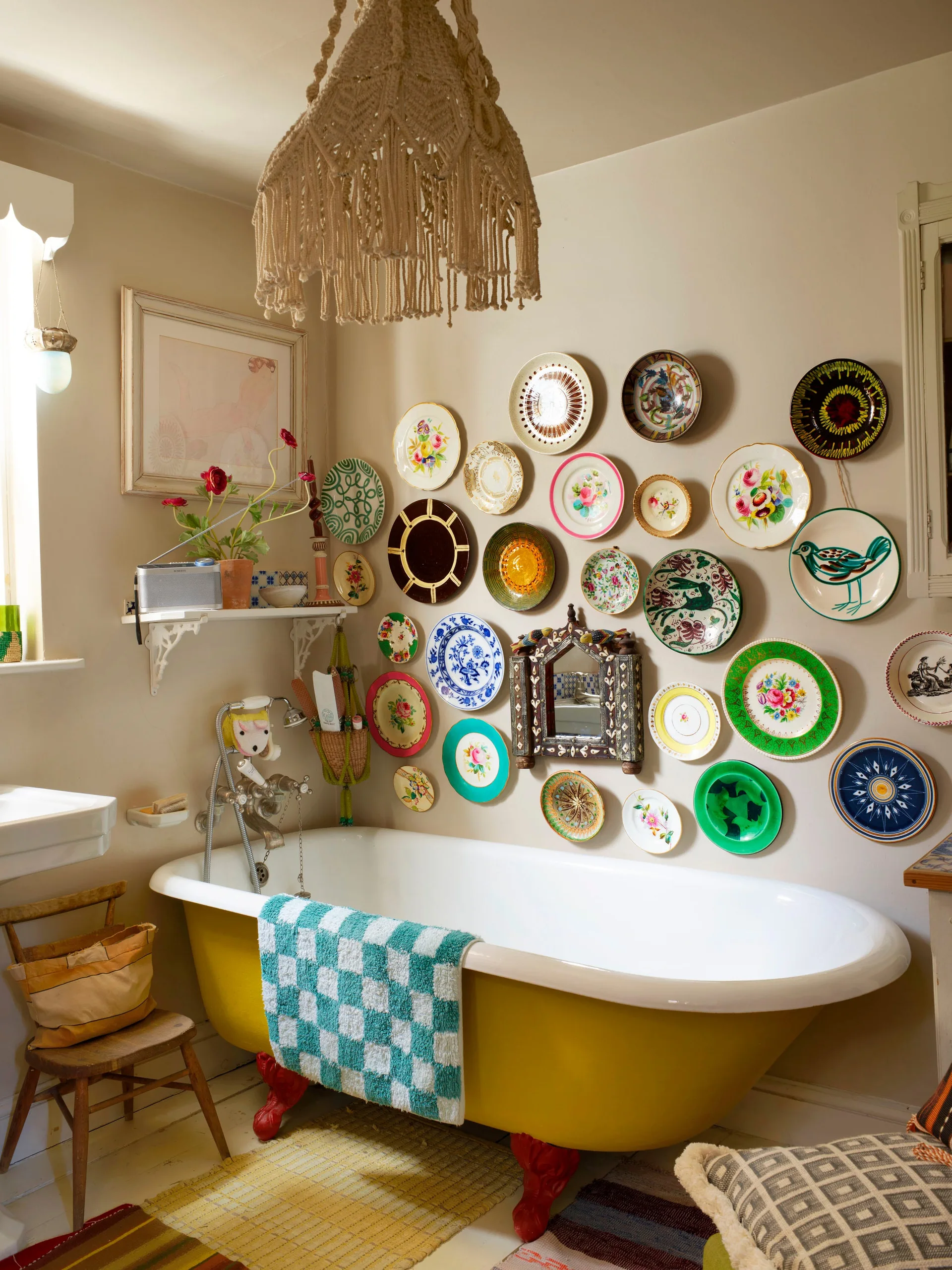 A yellow clawfoot bathtub sits against a beige wall adorned with a gallery of colorful decorative ceramic plates under a fringed macrame pendant light.