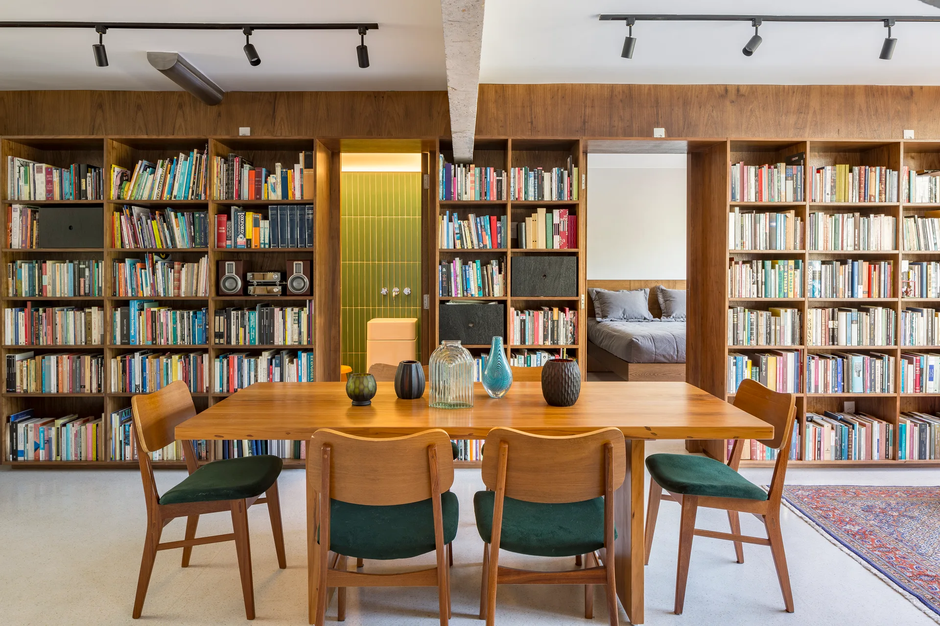 A wooden dining table and green upholstered chairs stand before a wall of built-in bookshelves containing doorways to adjacent rooms.