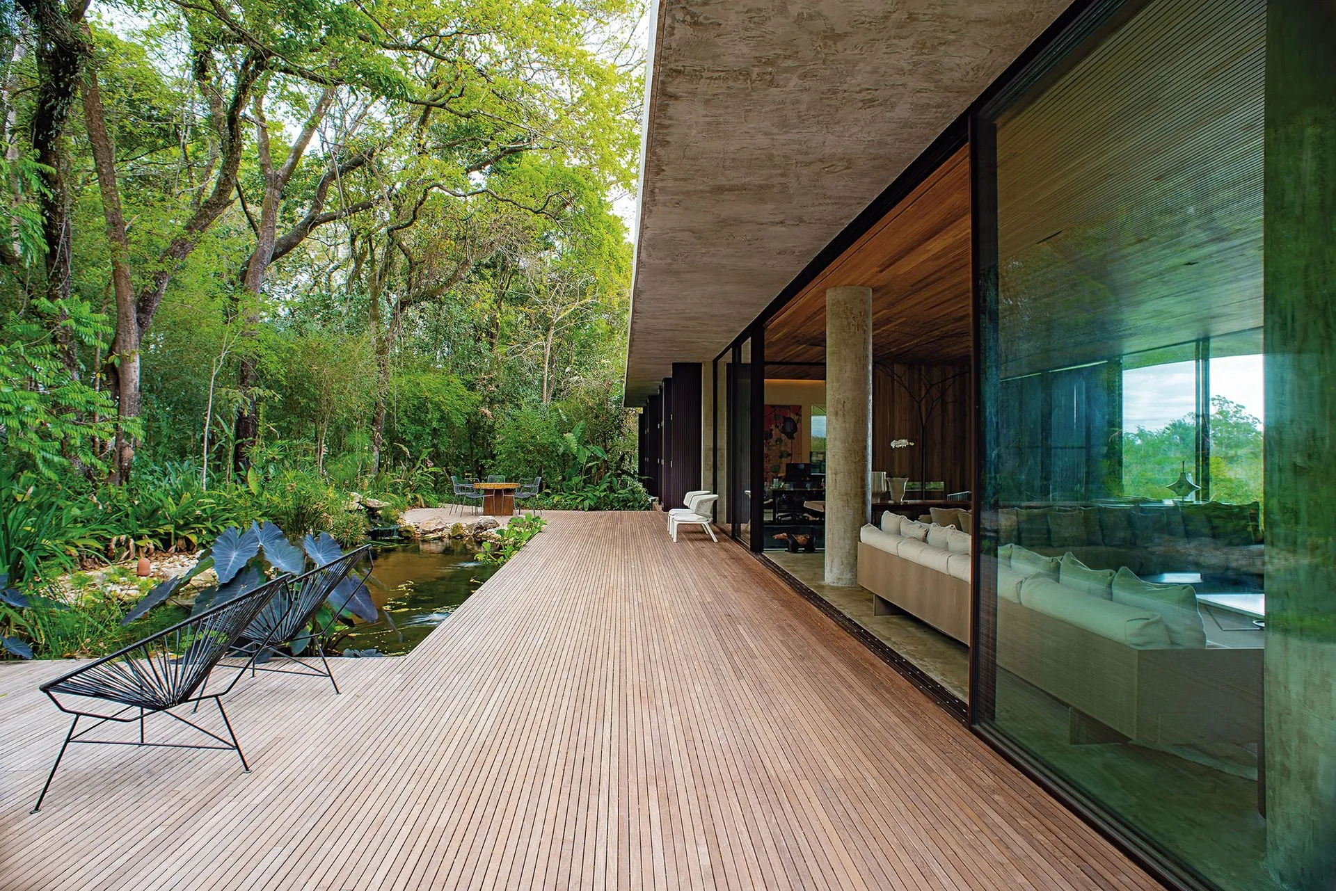 A spacious wooden deck with wire lounge chairs overlooks a garden pond next to a modern home with floor-to-ceiling glass walls.