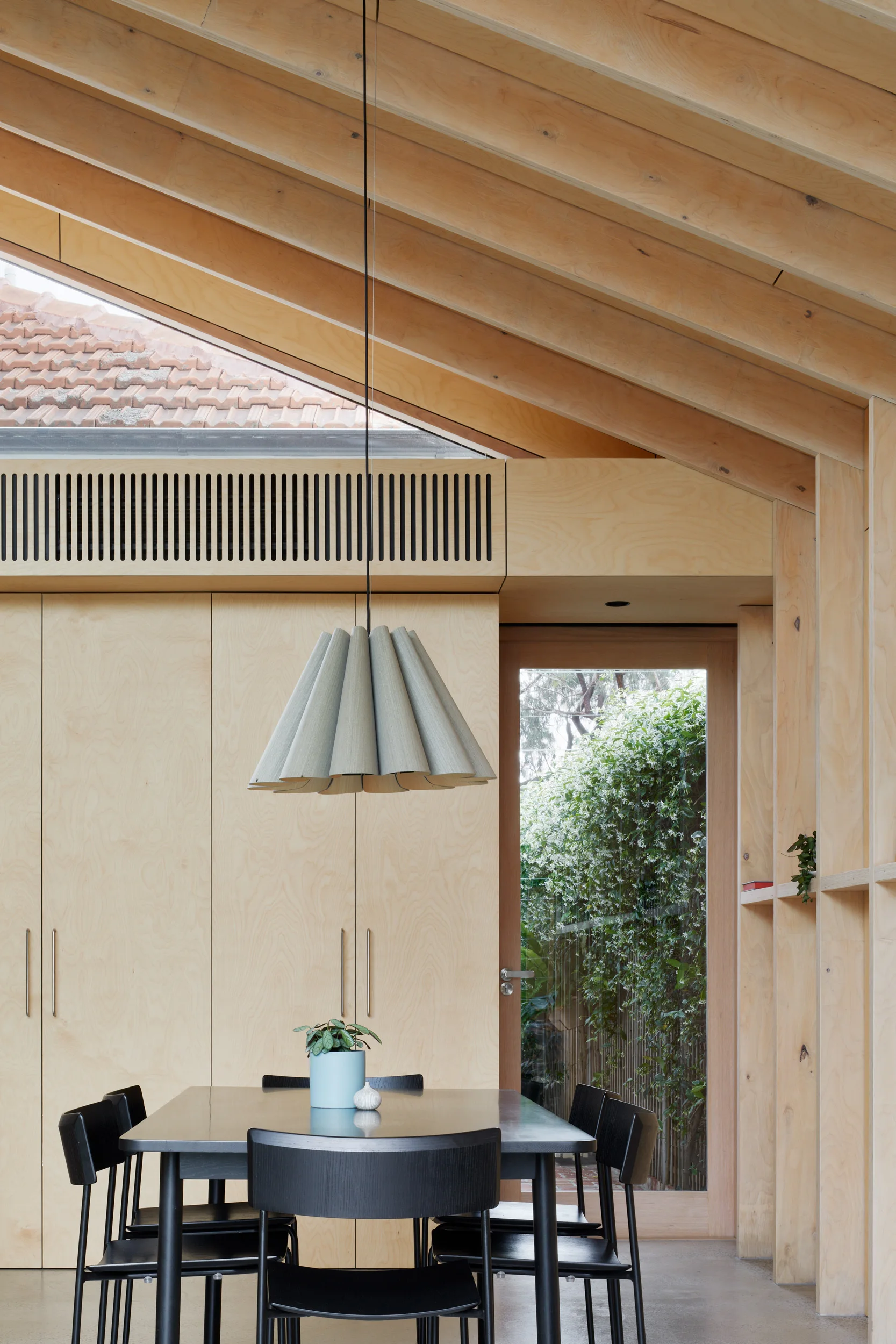A modern dining room features light plywood walls and ceiling beams, a black dining table with matching chairs, and a pleated hanging pendant light.