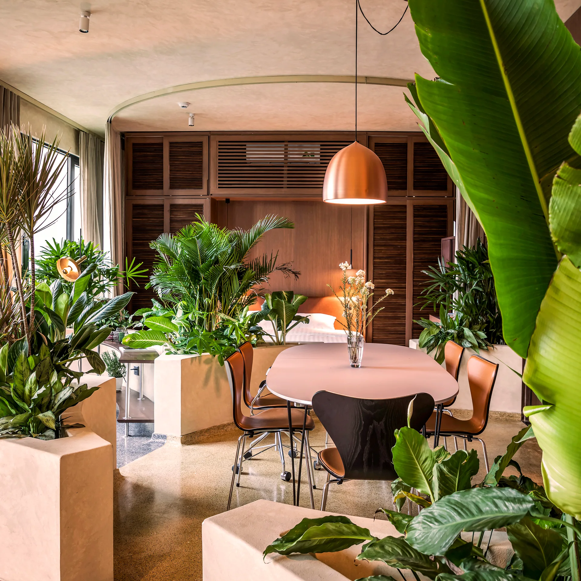 An oval dining table with mid-century style chairs is surrounded by custom plaster planters filled with lush green tropical plants.