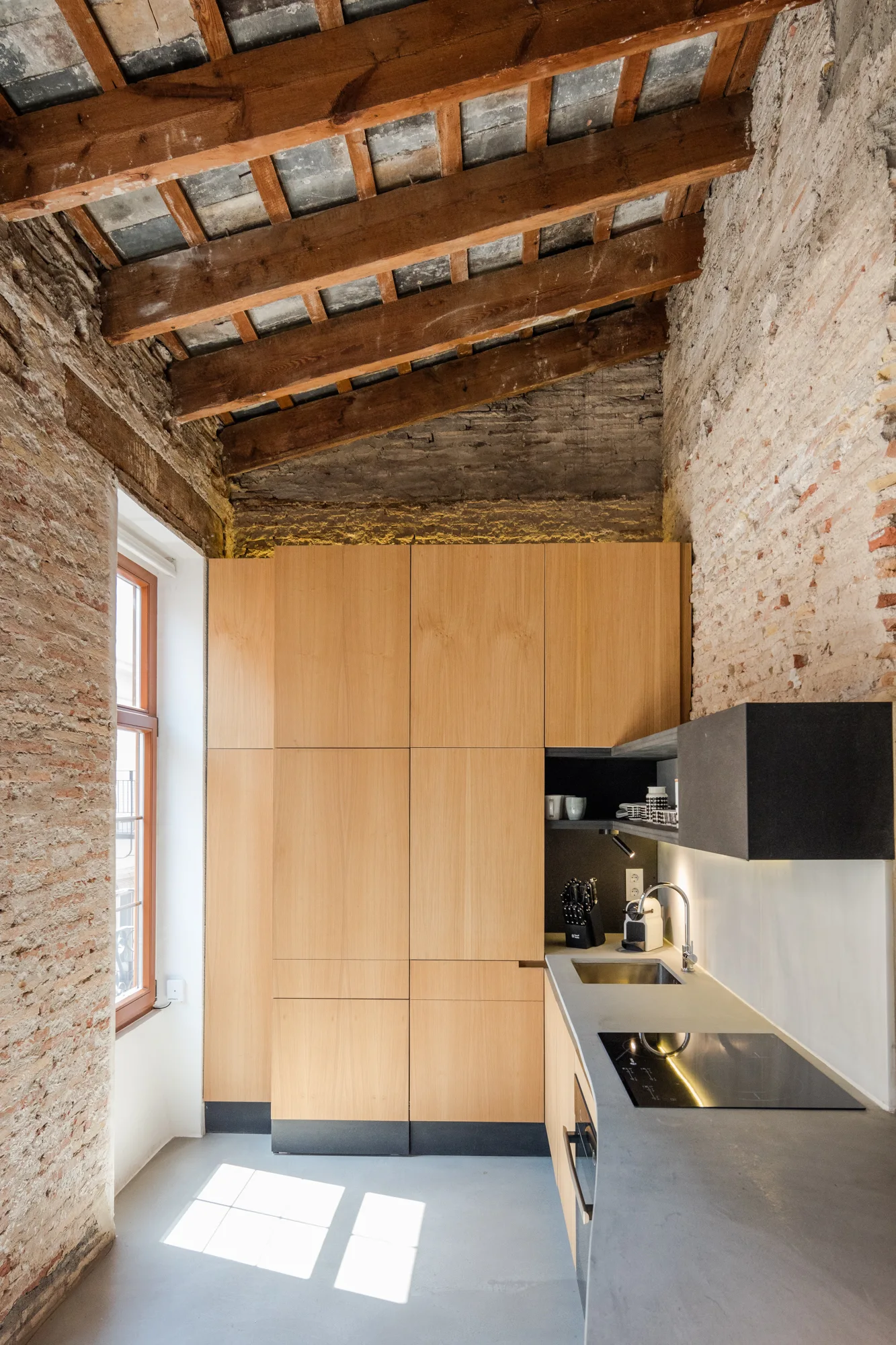 A modern kitchen features minimalist light wood cabinetry, a grey countertop, raw exposed brick walls, and rustic timber ceiling beams.