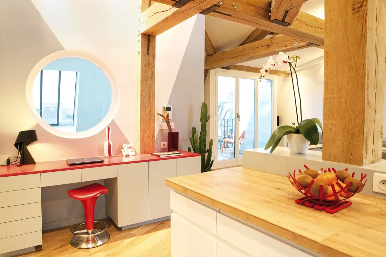 A bright interior space features a built-in grey desk with a red top, a circular mirror, and thick rustic wood beams overhead.