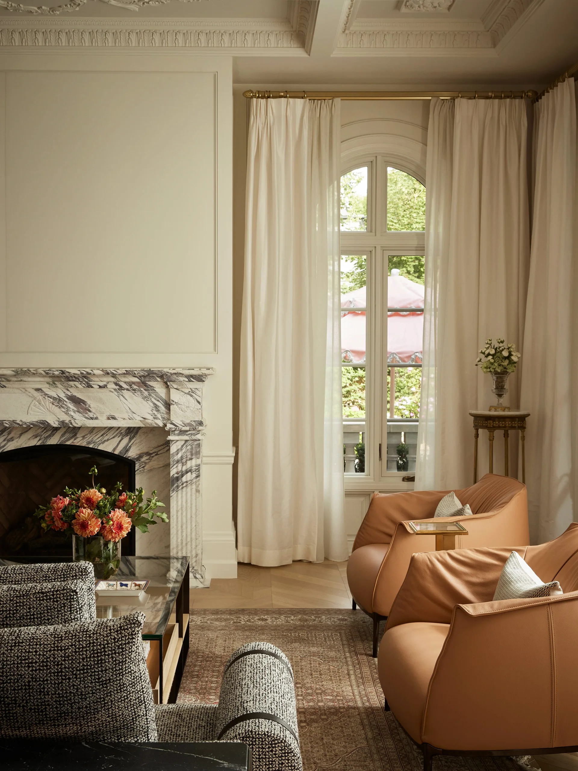 A living room features a highly veined marble fireplace, two tan leather armchairs, and floor-length white curtains framing an arched window.