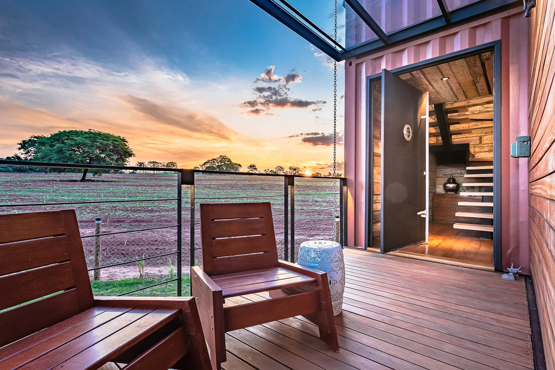 A wooden deck on a container home features two wooden armchairs, a patterned ceramic stool, and a cable railing overlooking a sunset.