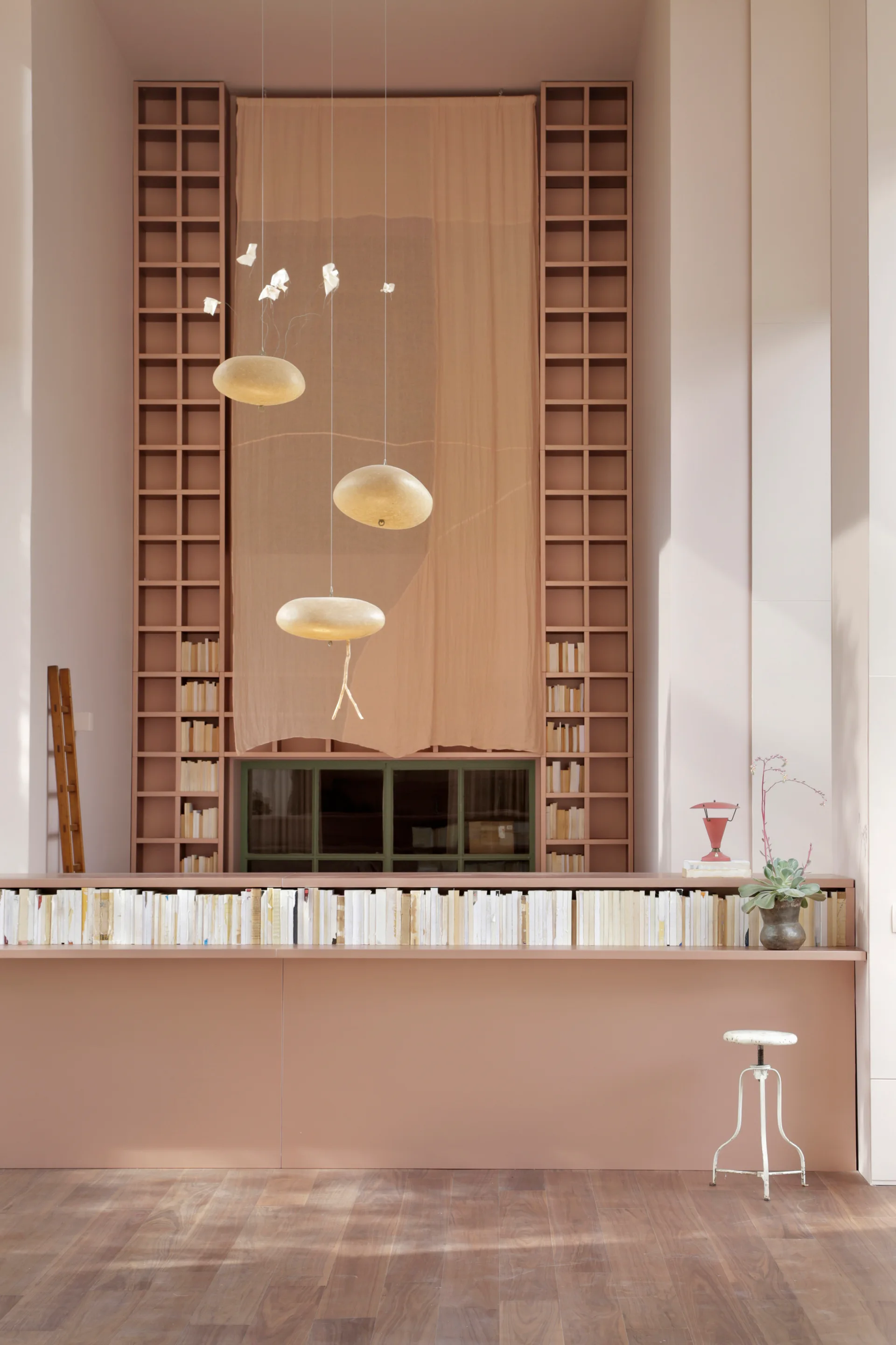 Symmetrical terracotta shelving frames a central draped backdrop with three suspended oval pendant lights above a long, book-lined counter.