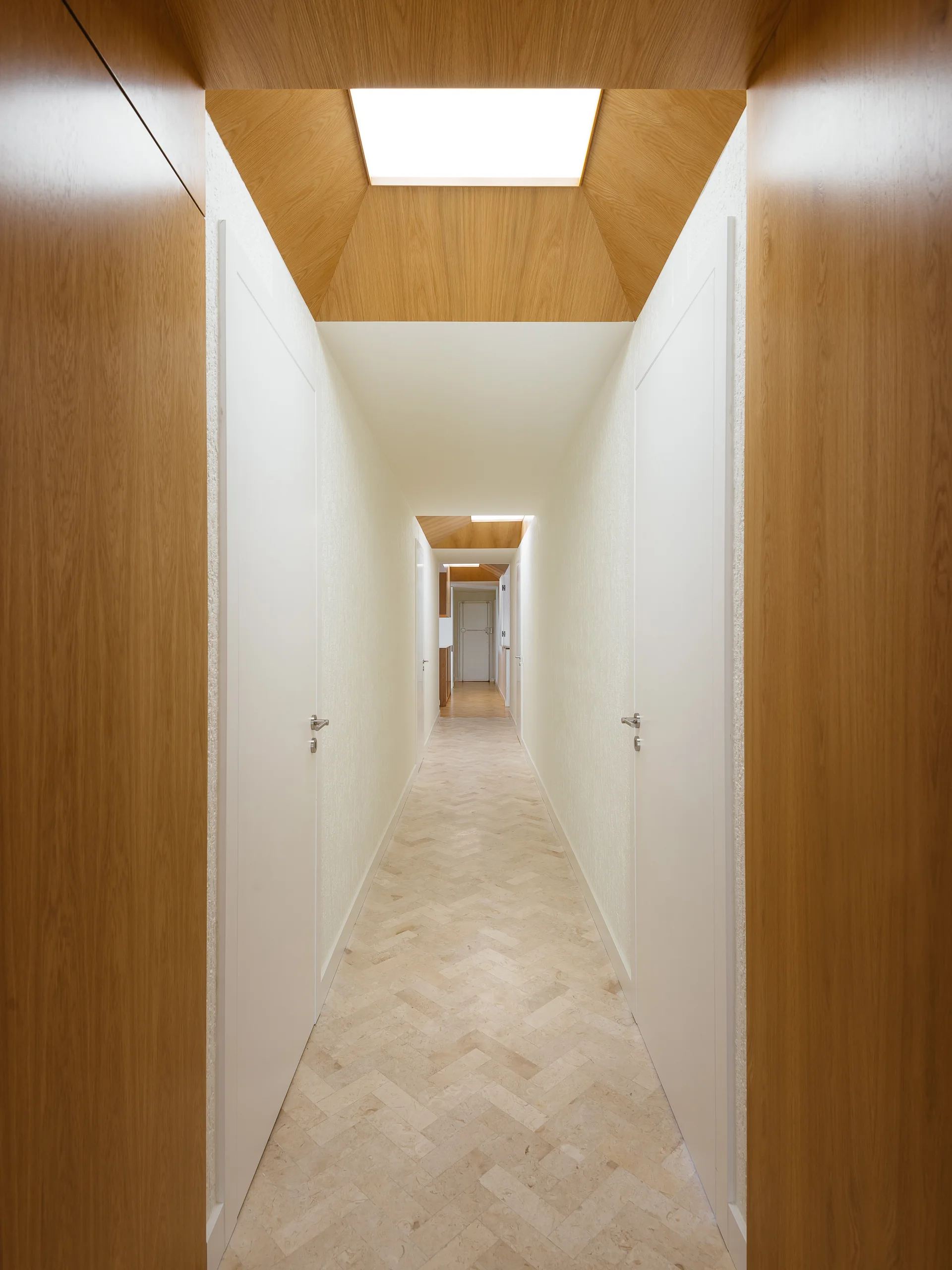 A long, minimalist hallway featuring beige herringbone stone flooring, white walls, and a recessed skylight surrounded by warm wood paneling.