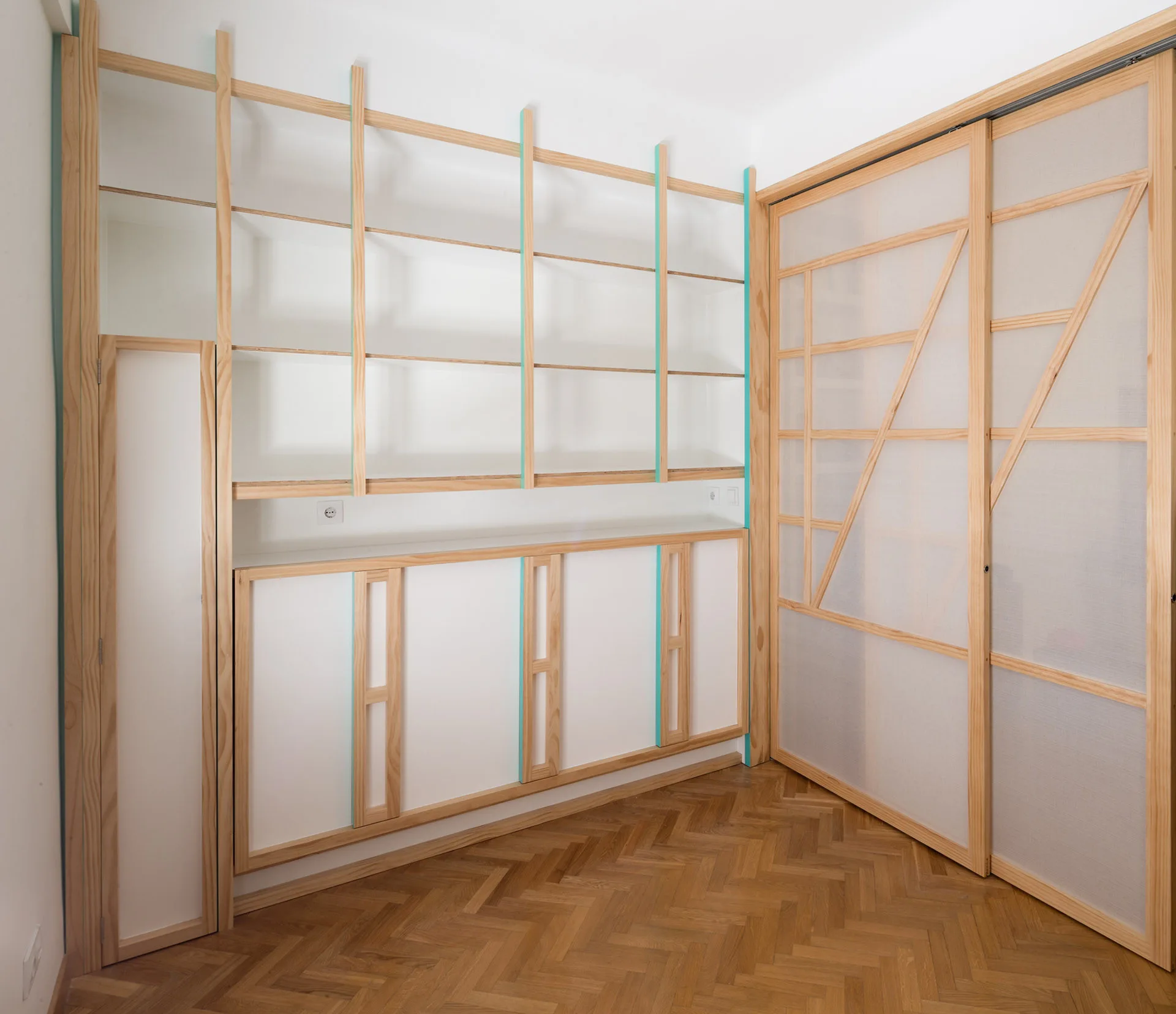 Custom light wood built-in shelving and cabinets with thin teal edges sit next to sliding closet doors with geometric patterns on herringbone parquet flooring.