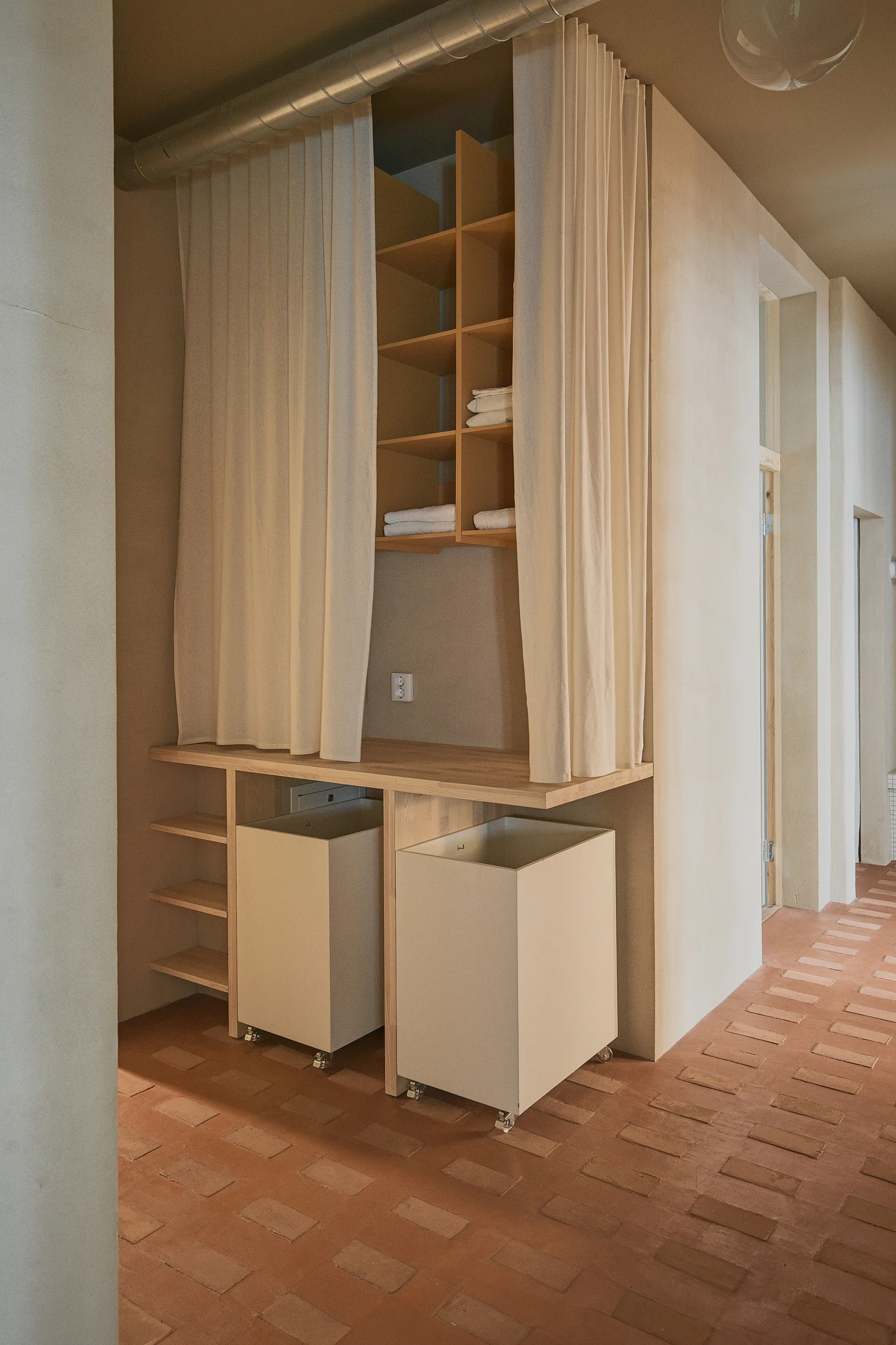 A minimalist laundry station features light wood shelving framed by beige curtains, two rolling hampers, and terracotta tile flooring.