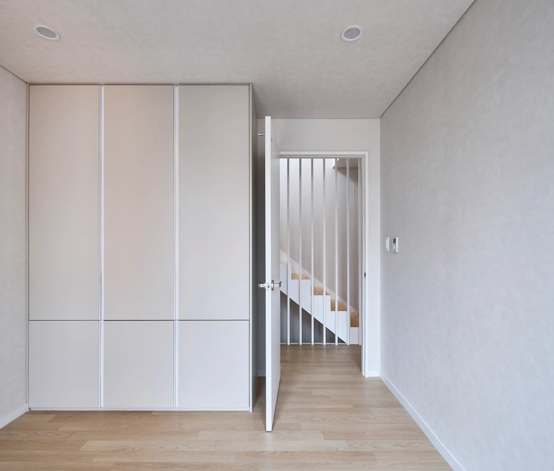 A minimalist hallway features a floor-to-ceiling beige built-in closet, light wood flooring, and an open door showing a white staircase with vertical rails.
