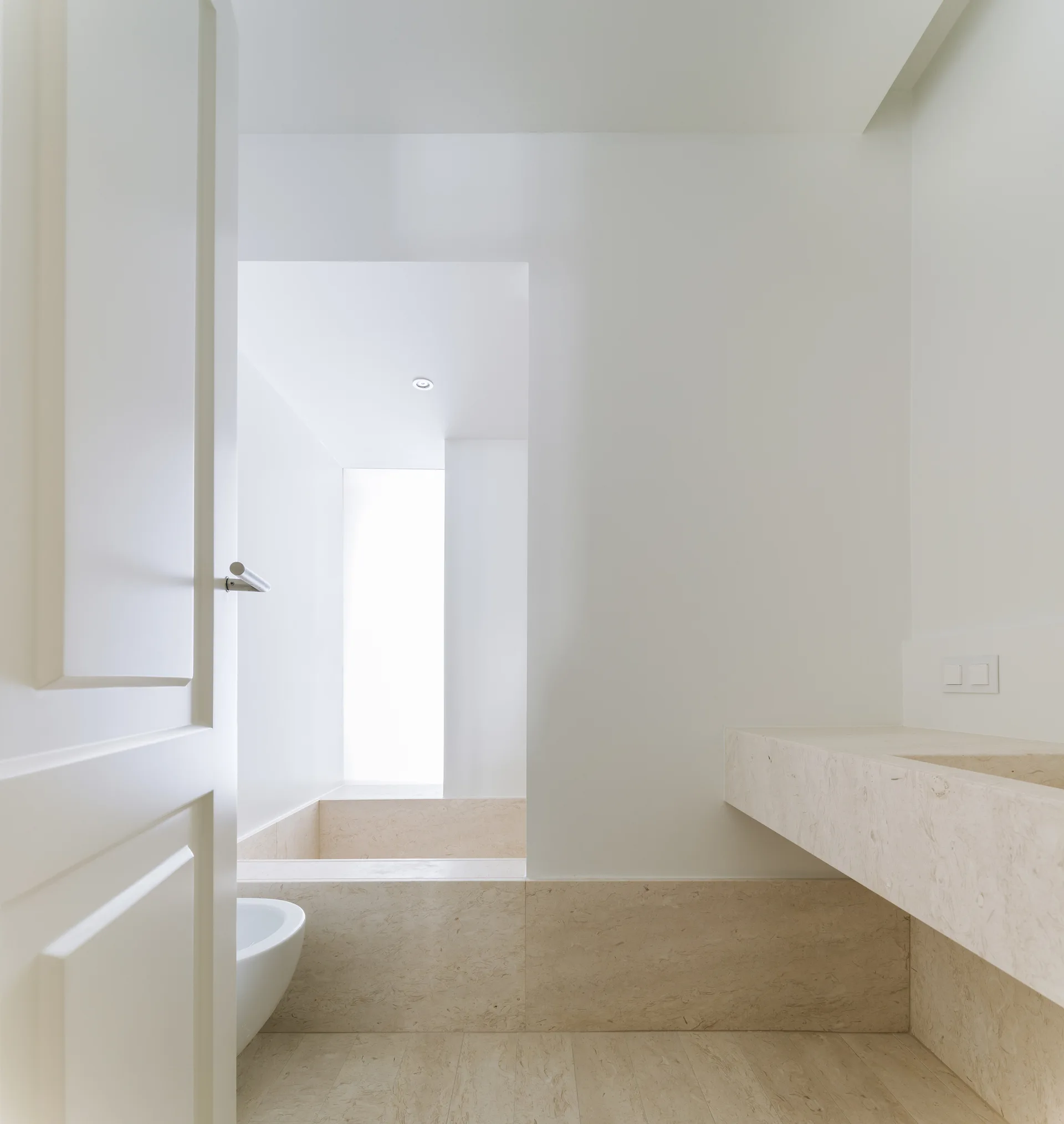 A minimalist bathroom featuring a floating beige stone vanity, matching stone floor, and a recessed built-in bathtub surrounded by white walls.