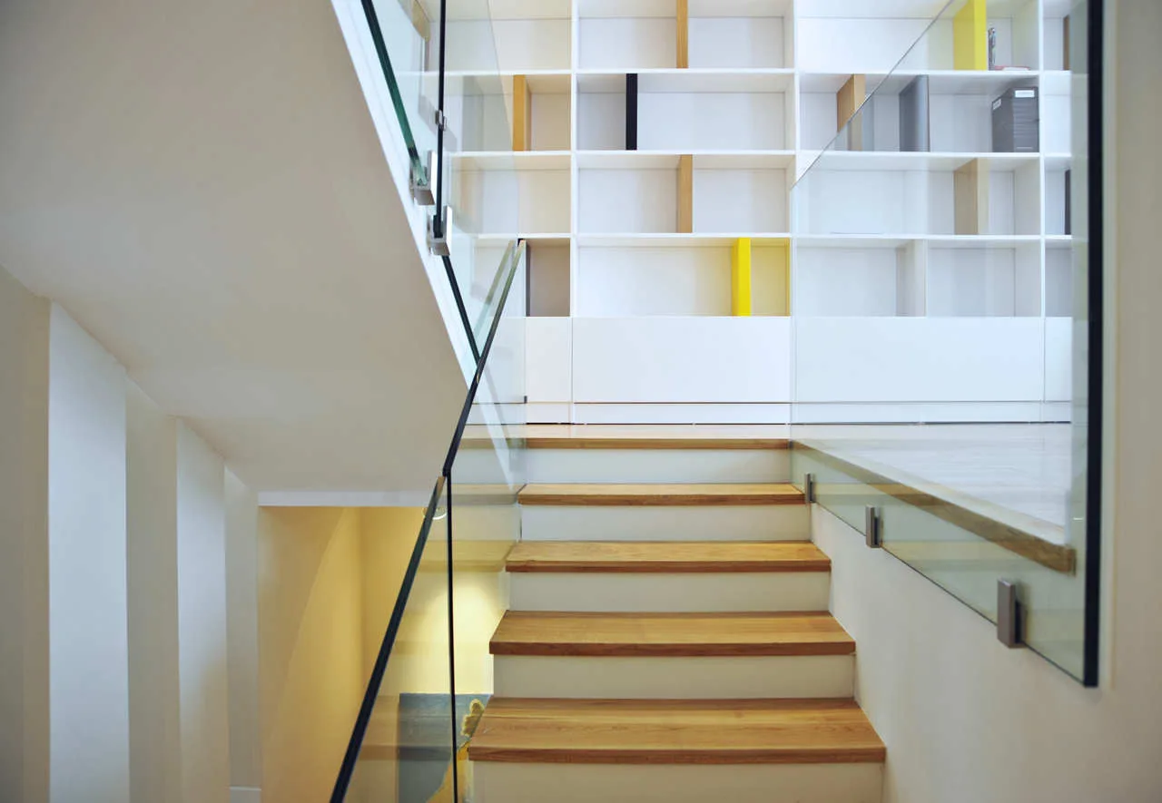 A modern staircase with light wood treads and a clear glass railing leads toward a large white built-in bookshelf.