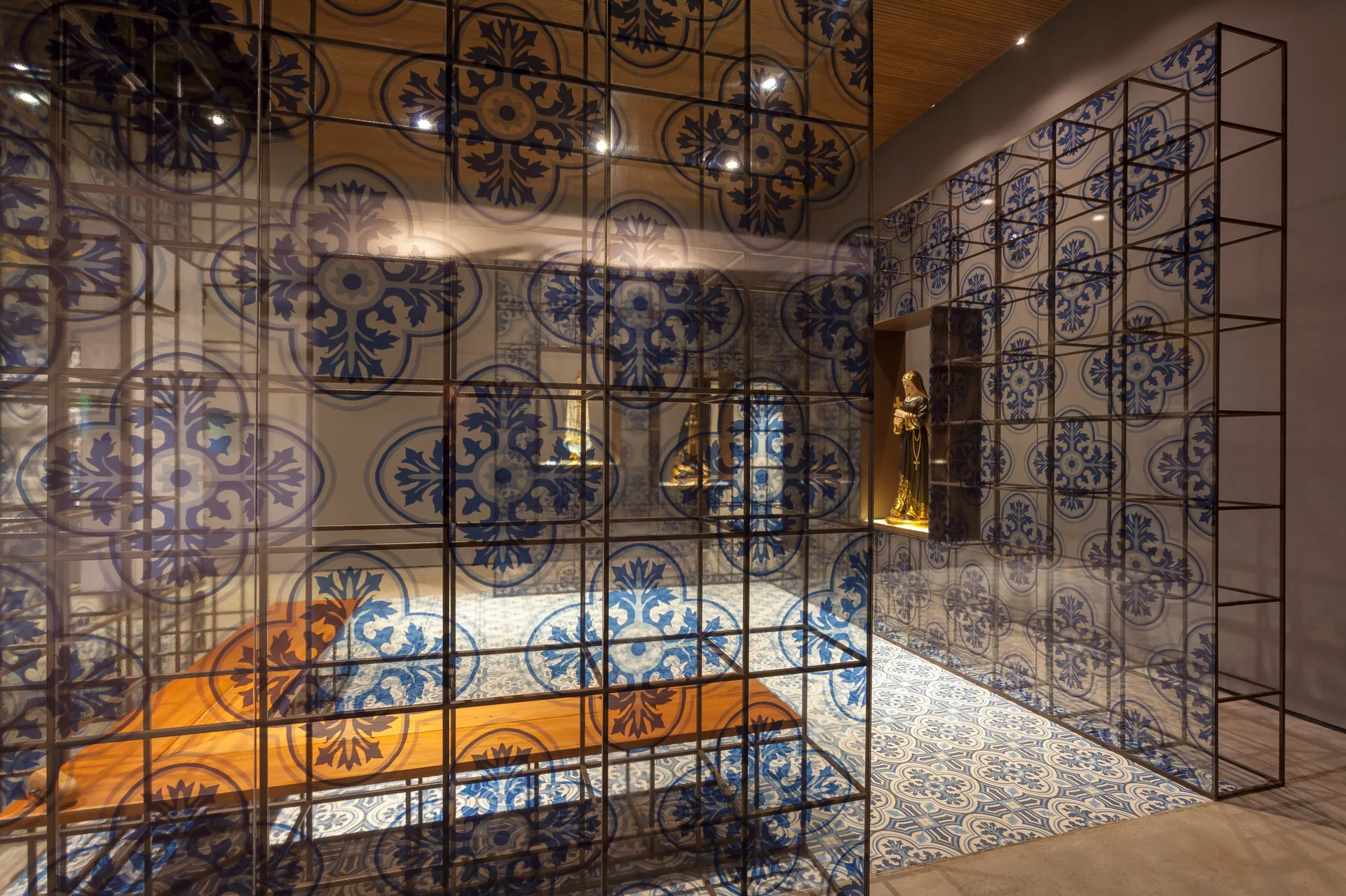 A structural metal grid partition with blue patterned glass panels stands on matching ornate tile flooring next to a lit wall niche.