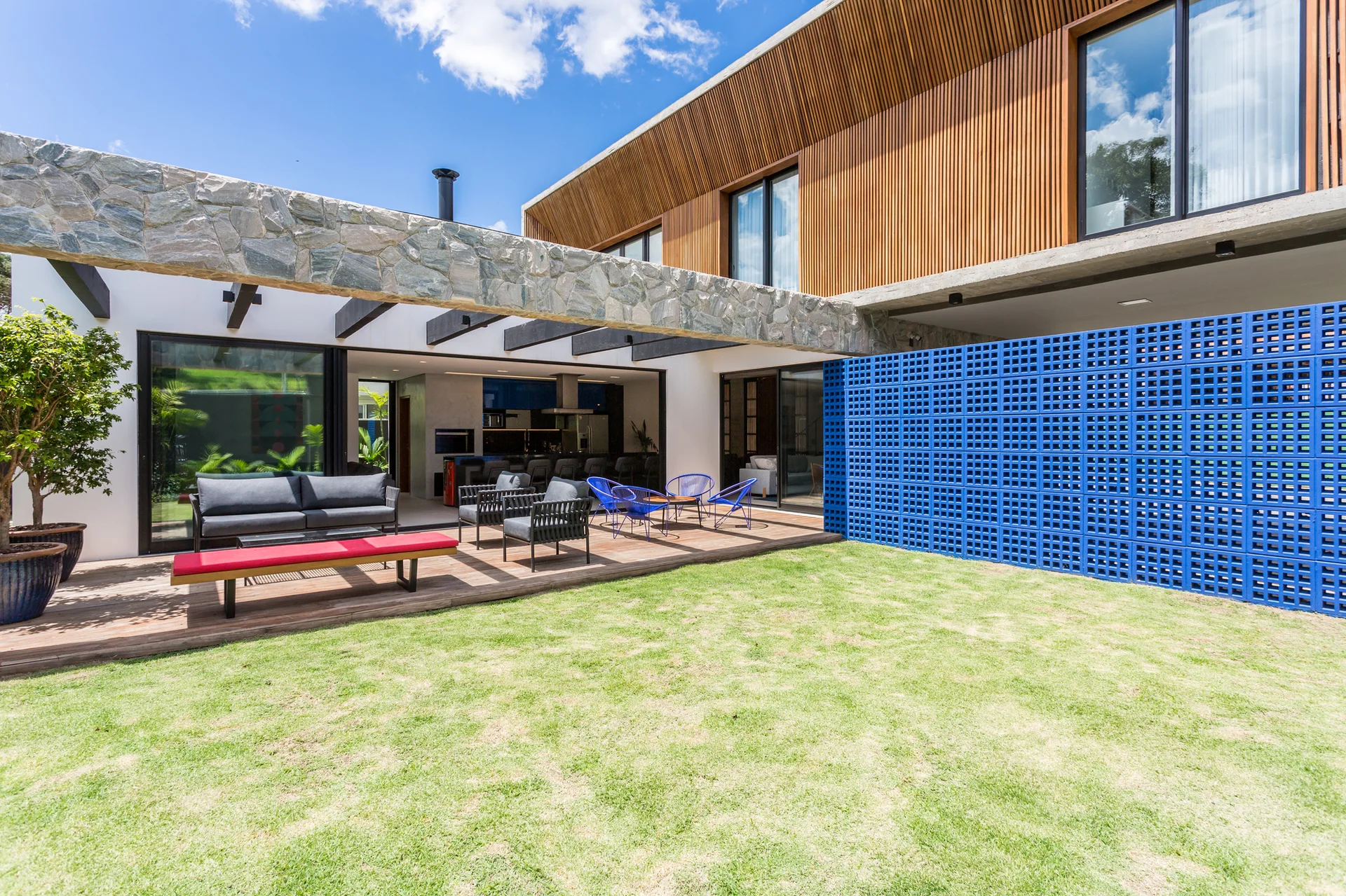 A modern outdoor patio featuring a wooden deck with lounge seating, a stone beam, and a vibrant blue perforated brick wall next to a green lawn.