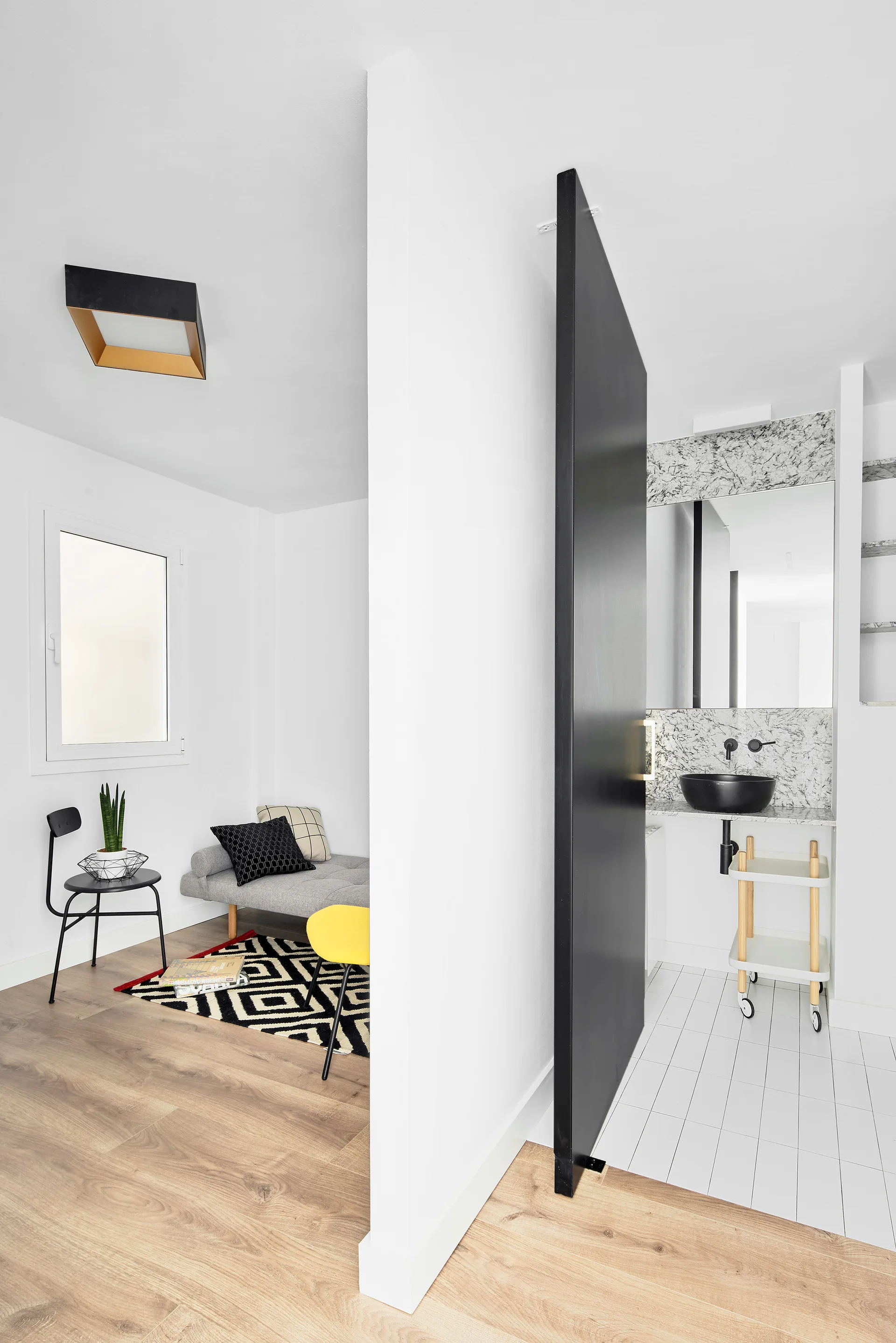 A white partition wall separates a small living corner with a grey daybed from a modern bathroom featuring a black pivot door and white tiled floor.