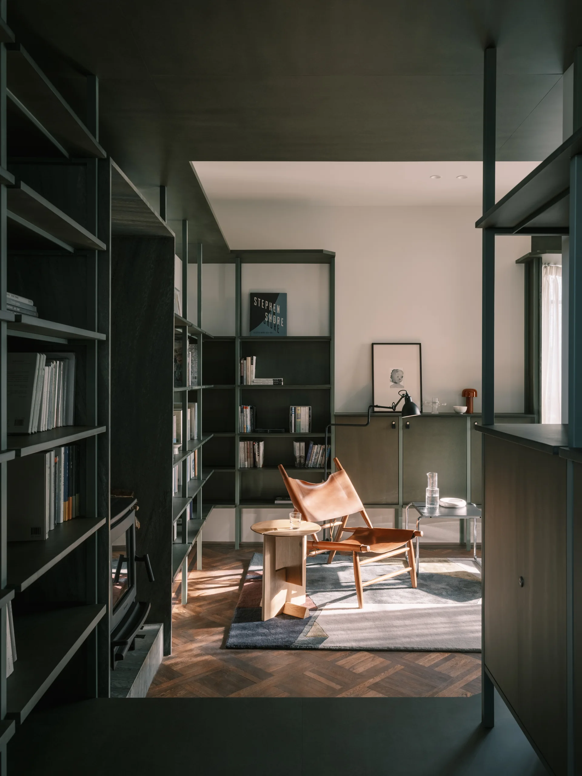A tan leather sling lounge chair sits on a geometric rug in a home library with deep green floor-to-ceiling bookcases.