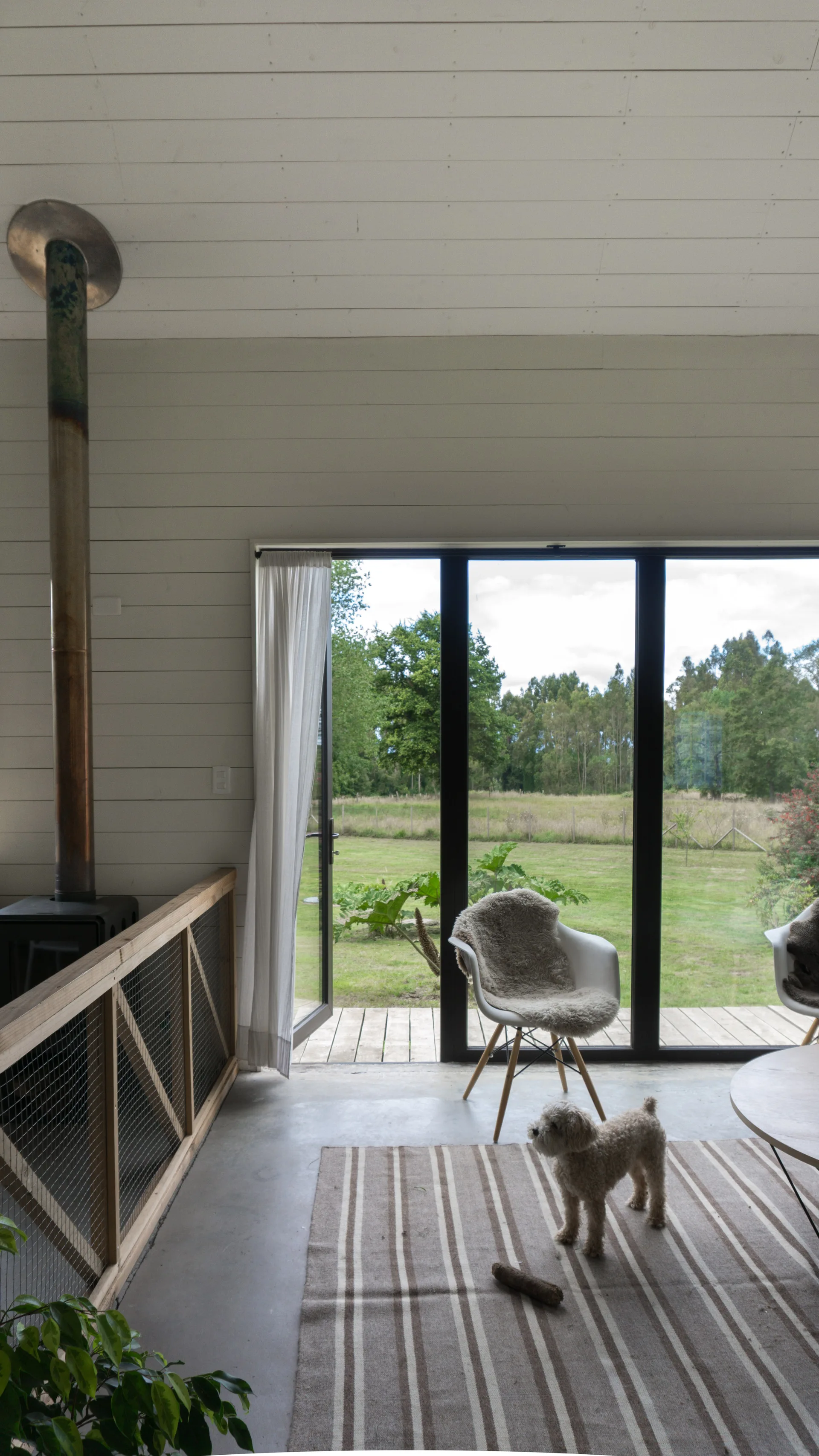 A modern rustic living room features a wood burning stove, a wire mesh railing, white paneled walls, and a large sliding glass door opening to a grassy field.