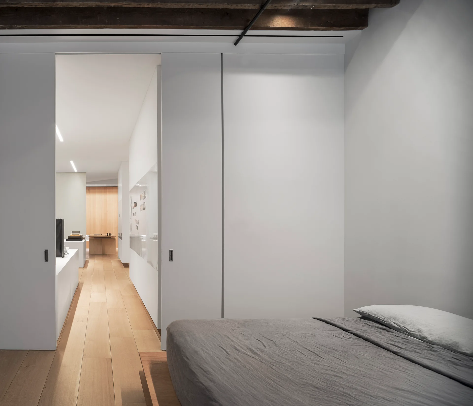 A minimalist bedroom with grey bedding and a large white sliding pocket door opening to an adjoining hallway with light wood floors.
