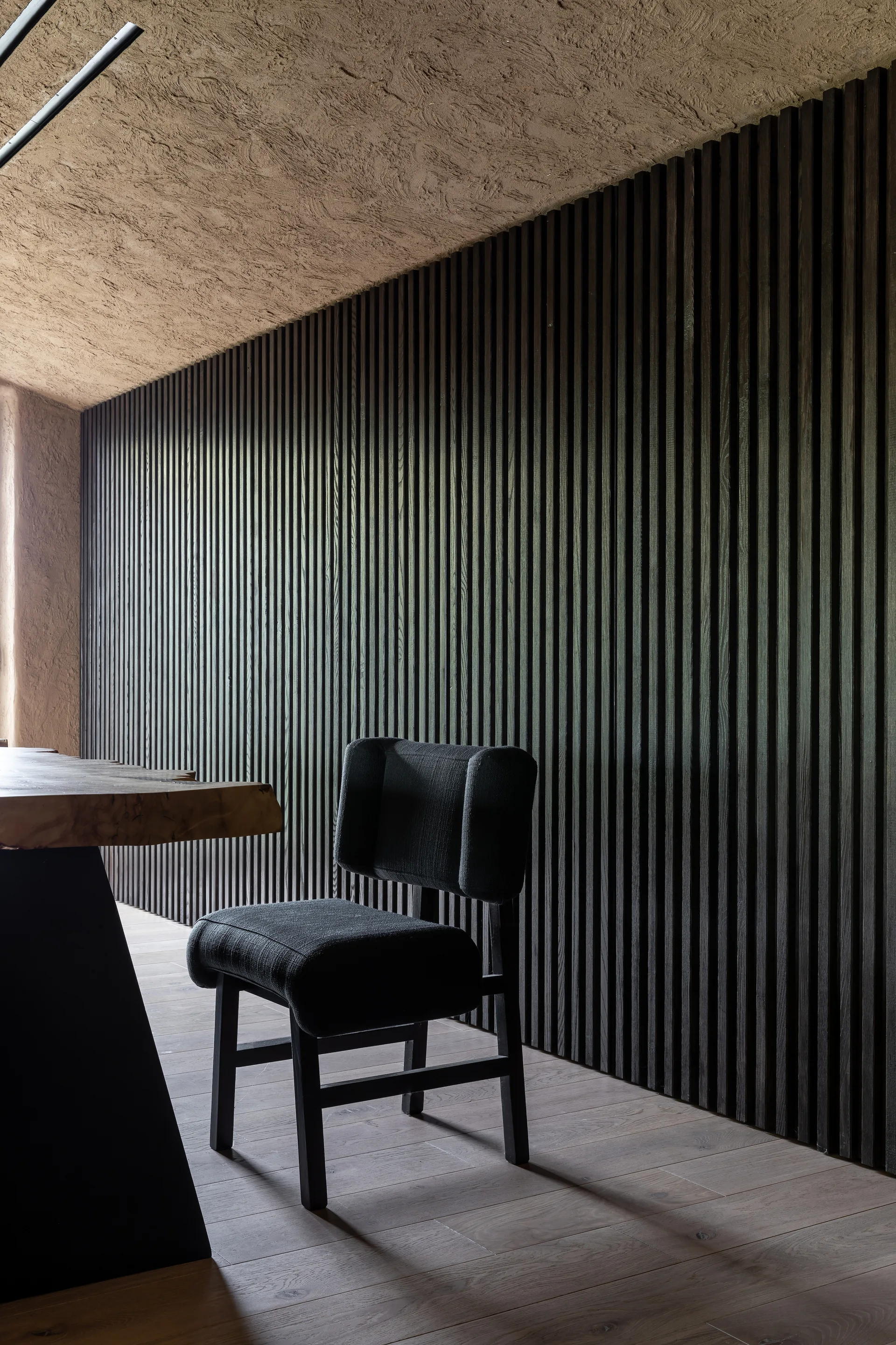 A charcoal upholstered dining chair sits next to a live-edge wooden table in front of a dark vertical slatted wood wall.