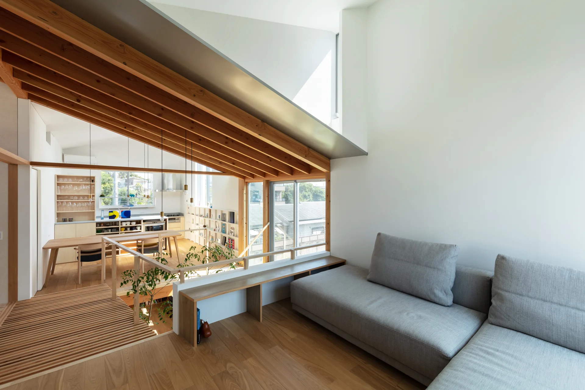 A modern split-level living room features a gray sofa, light wood floors, a low wooden railing, and a sloped ceiling with exposed wooden rafters.