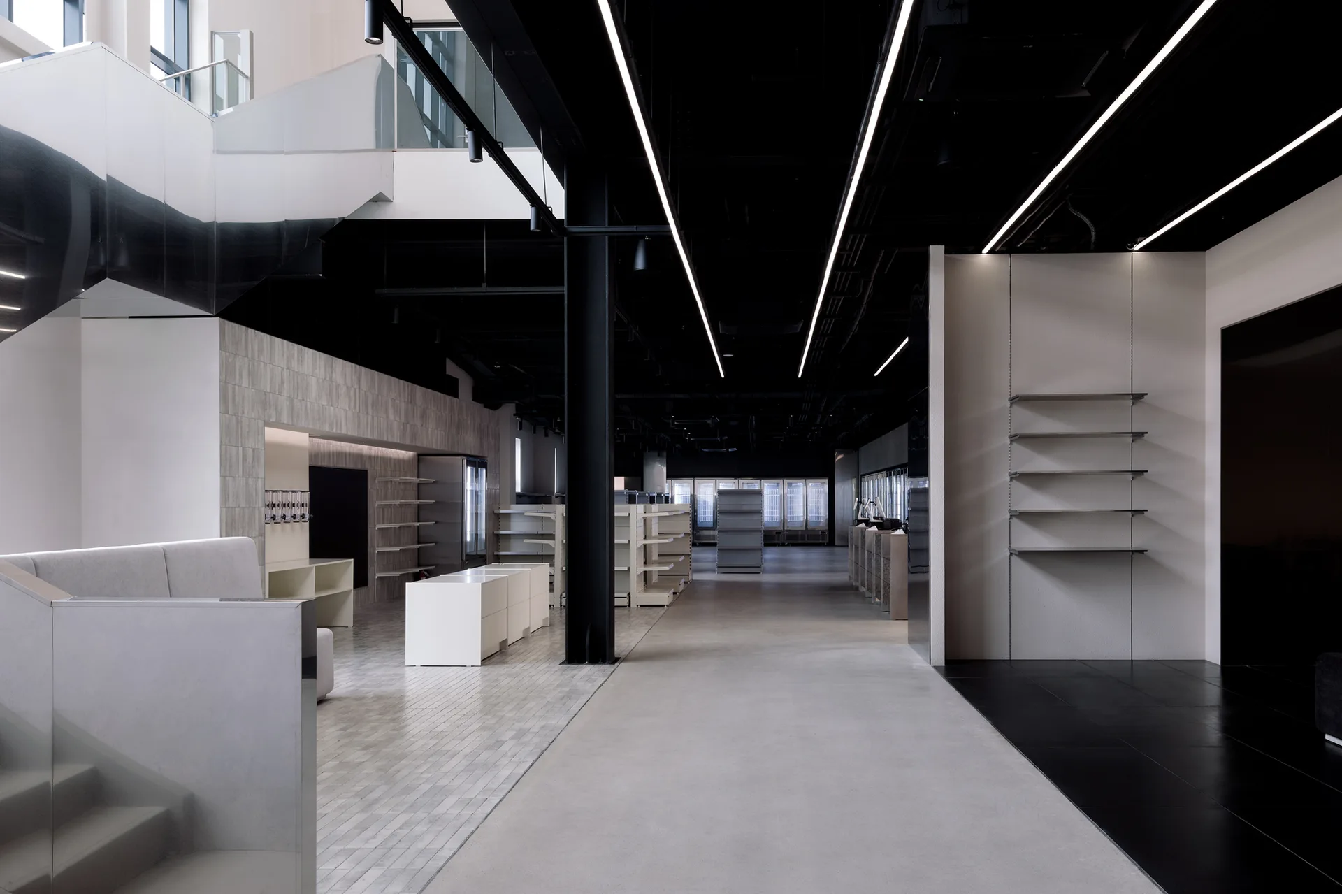 A minimalist commercial retail space features an exposed black ceiling with linear light fixtures, concrete flooring, and empty wall-mounted shelving.