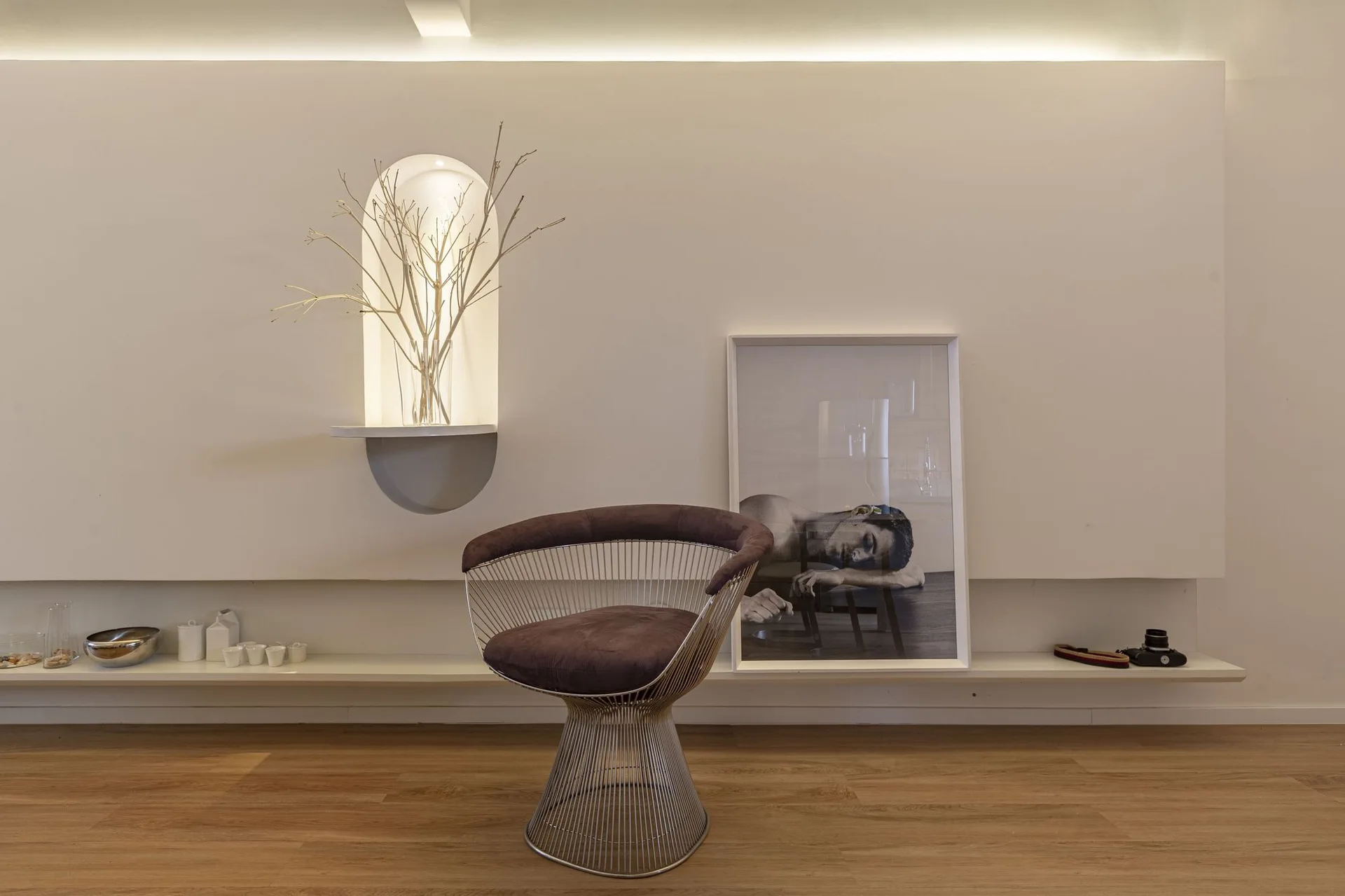 A sculptural wire-frame armchair stands in front of a white wall with a glowing arched niche and a long low shelf.