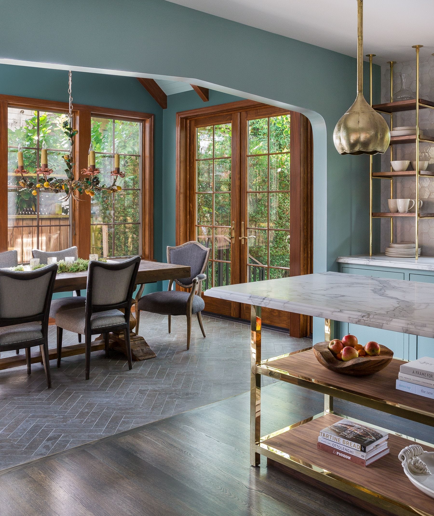 A dining room with teal blue walls, herringbone tile floors, a wooden table with upholstered chairs, and wood-framed French doors leading outside.