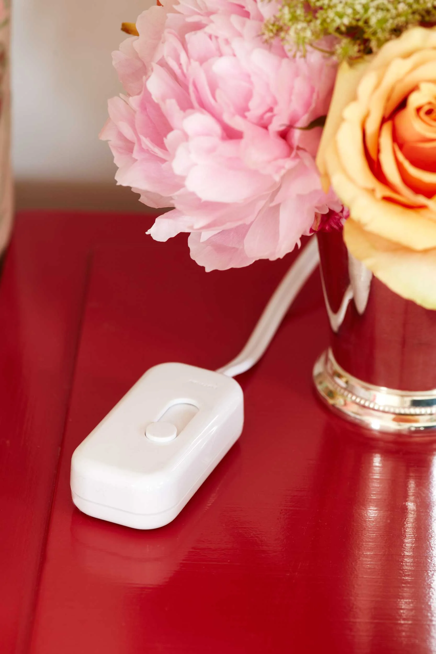 A close-up of a white plastic inline light switch resting on a glossy red wooden tabletop beside a silver vase of pink and yellow flowers.