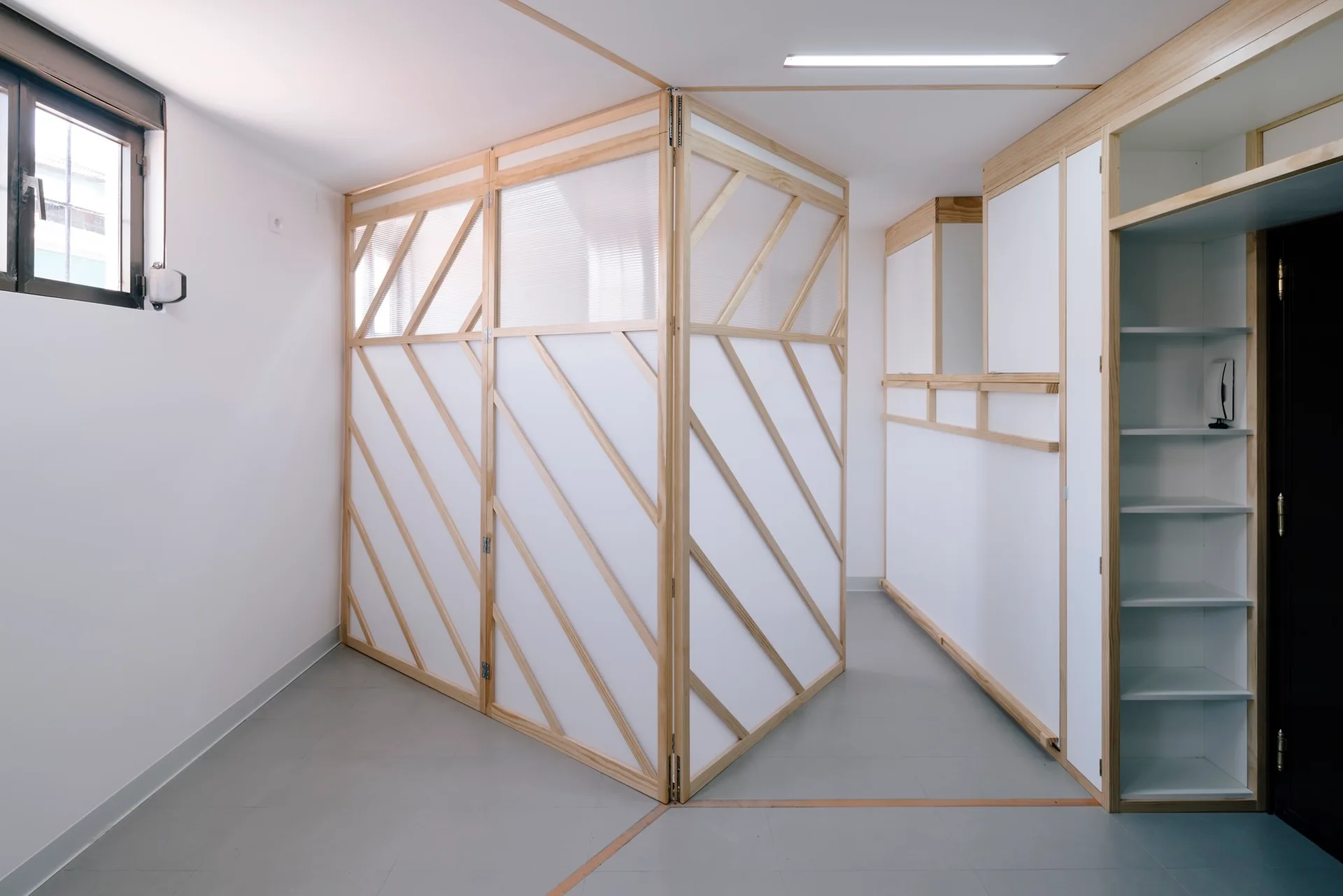 A light wood partition wall with diagonal slats and translucent panels divides a minimalist room with built-in shelving and light gray floors.
