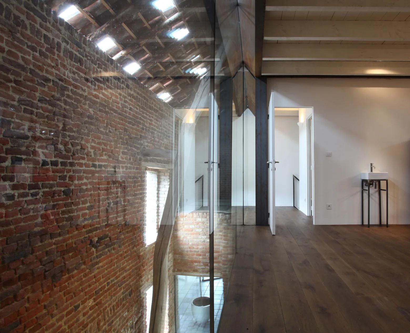 An upper landing with dark wood floors and white walls is separated from a double-height brick wall by a frameless glass partition.