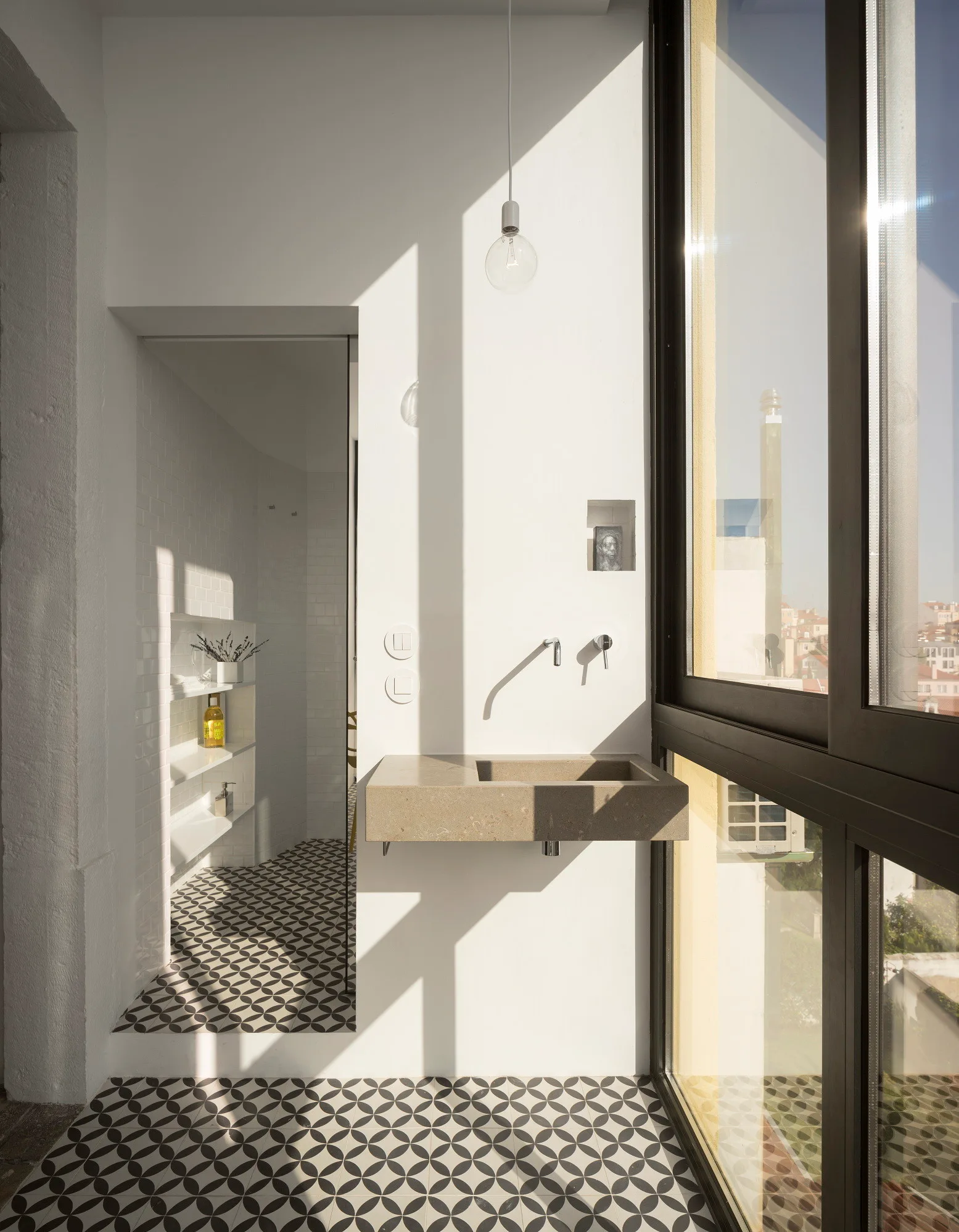A modern bathroom features a wall-mounted beige stone sink, a chrome faucet, black-and-white geometric floor tiles, and a large sunlit window.
