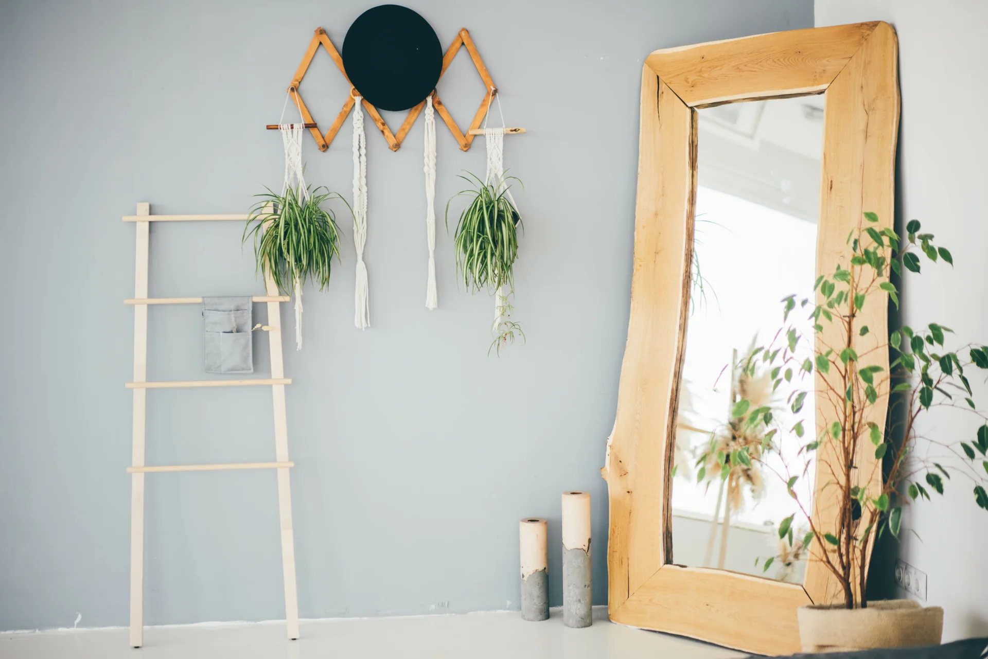 A large live-edge wooden floor mirror leans against a grey wall next to a decorative ladder and hanging macrame planters.