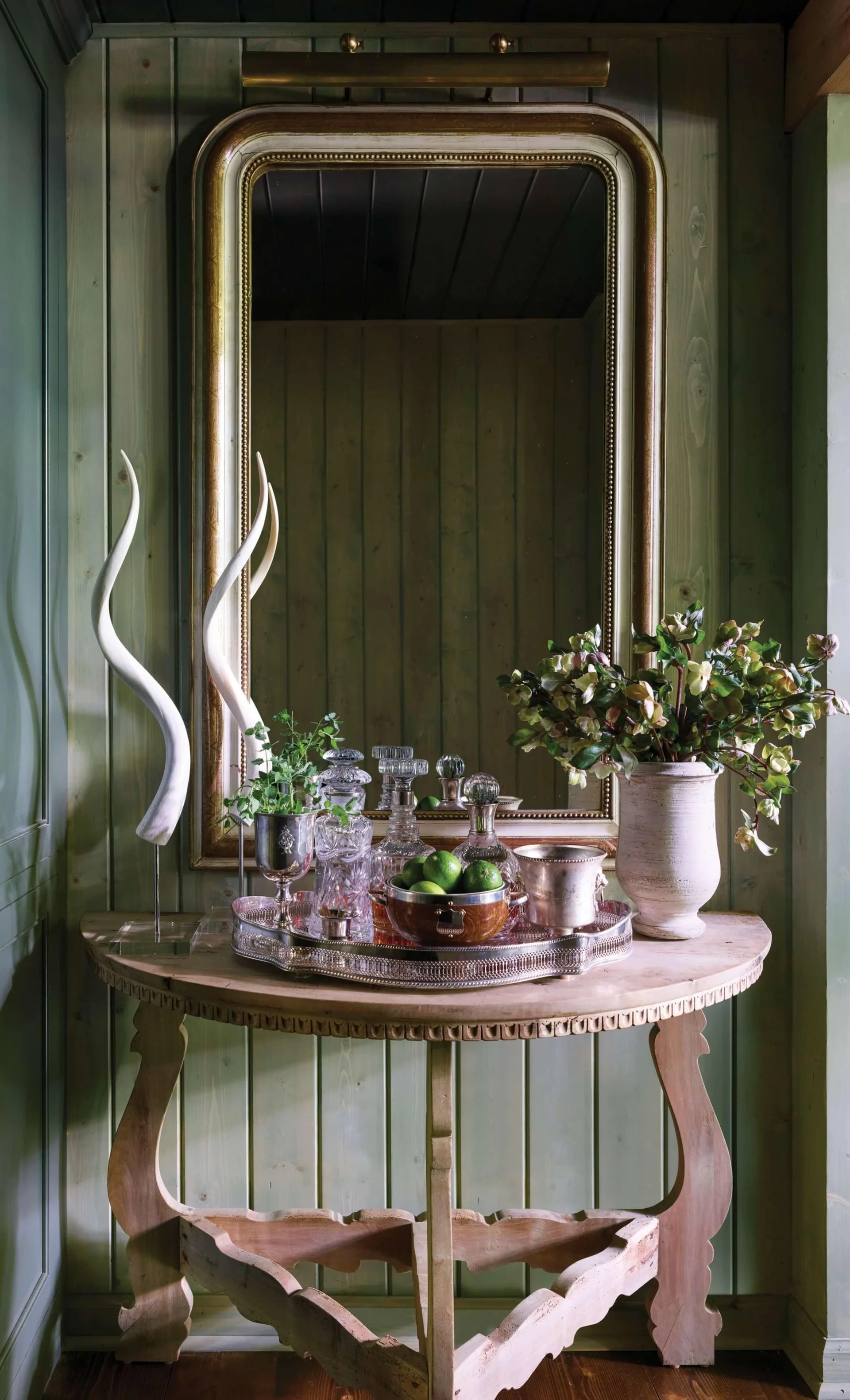 A semicircular wood console table styled with a silver bar tray, glass decanters, and a floral vase against sage green vertical wood paneling.