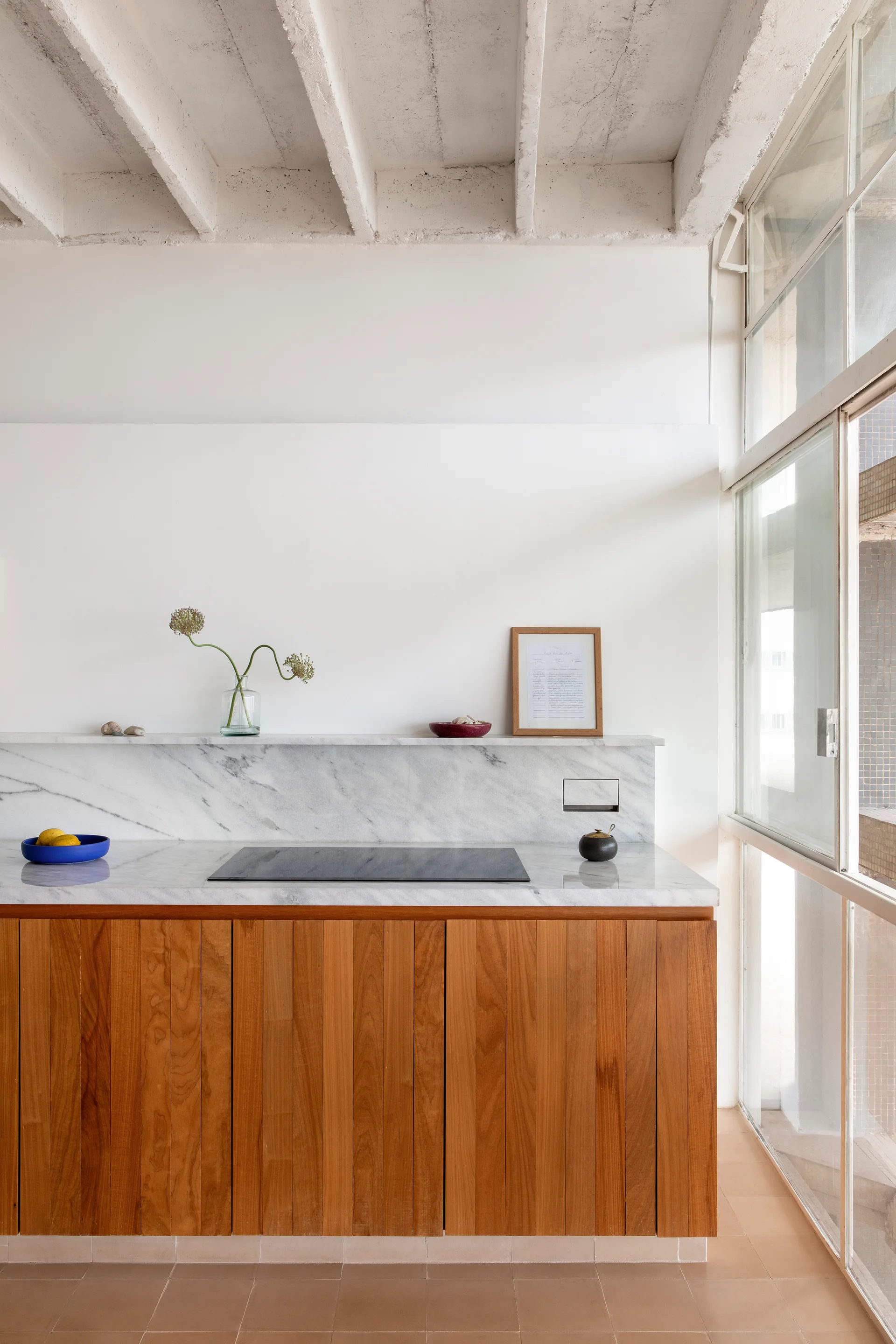 A minimalist kitchen features vertical wood cabinets, a white marble countertop with a matching ledge, and exposed painted concrete ceiling beams.