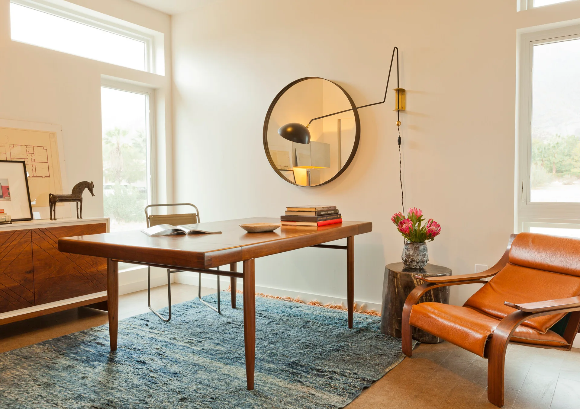 A bright home office featuring a mid-century modern wooden desk on a blue rug, a round wall mirror, and a tan leather lounge chair.