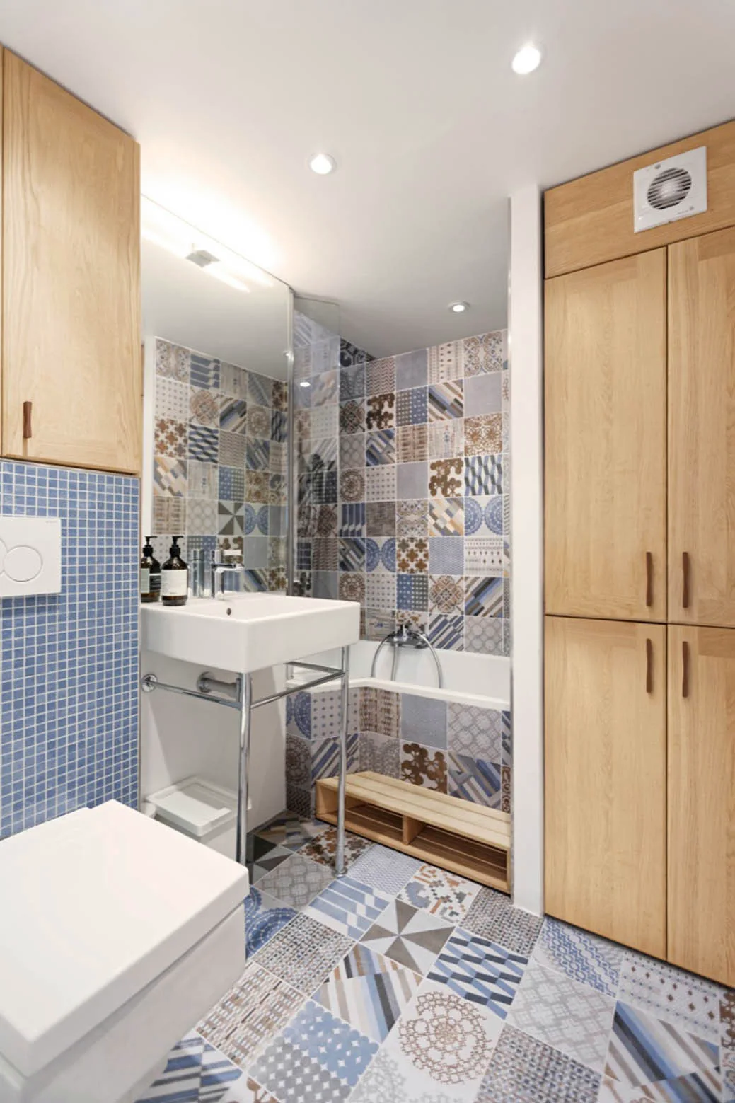 A modern bathroom features patchwork blue and white patterned tile on the floor and bathtub wall alongside light oak cabinetry and a metal frame sink.