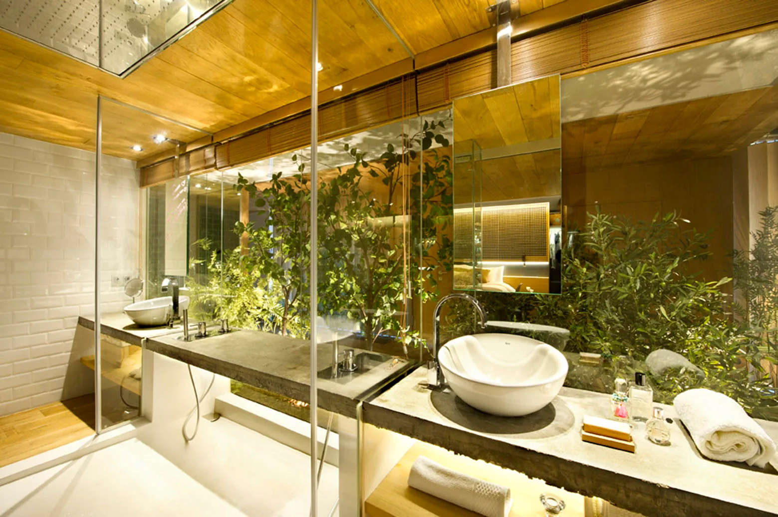 A modern bathroom features a grey concrete vanity with white vessel sinks next to a large glass wall showcasing green plants.