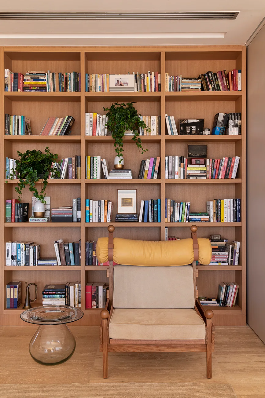A floor-to-ceiling wooden bookcase filled with books stands behind a mid-century armchair with a yellow bolster pillow and a clear glass side table.