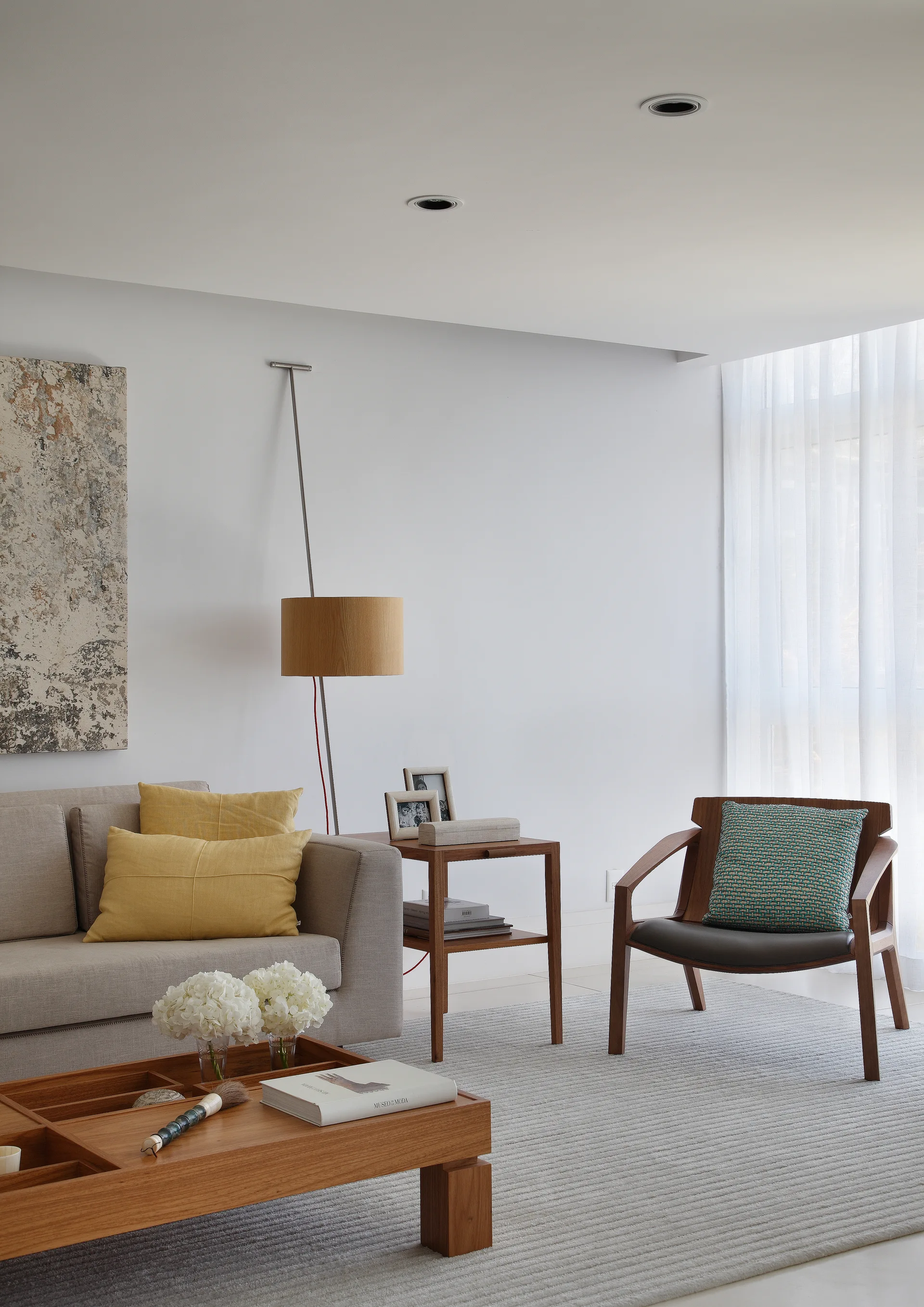 A modern living room features a light grey sofa with yellow pillows, a wooden coffee table, and a wooden armchair on a textured grey rug.