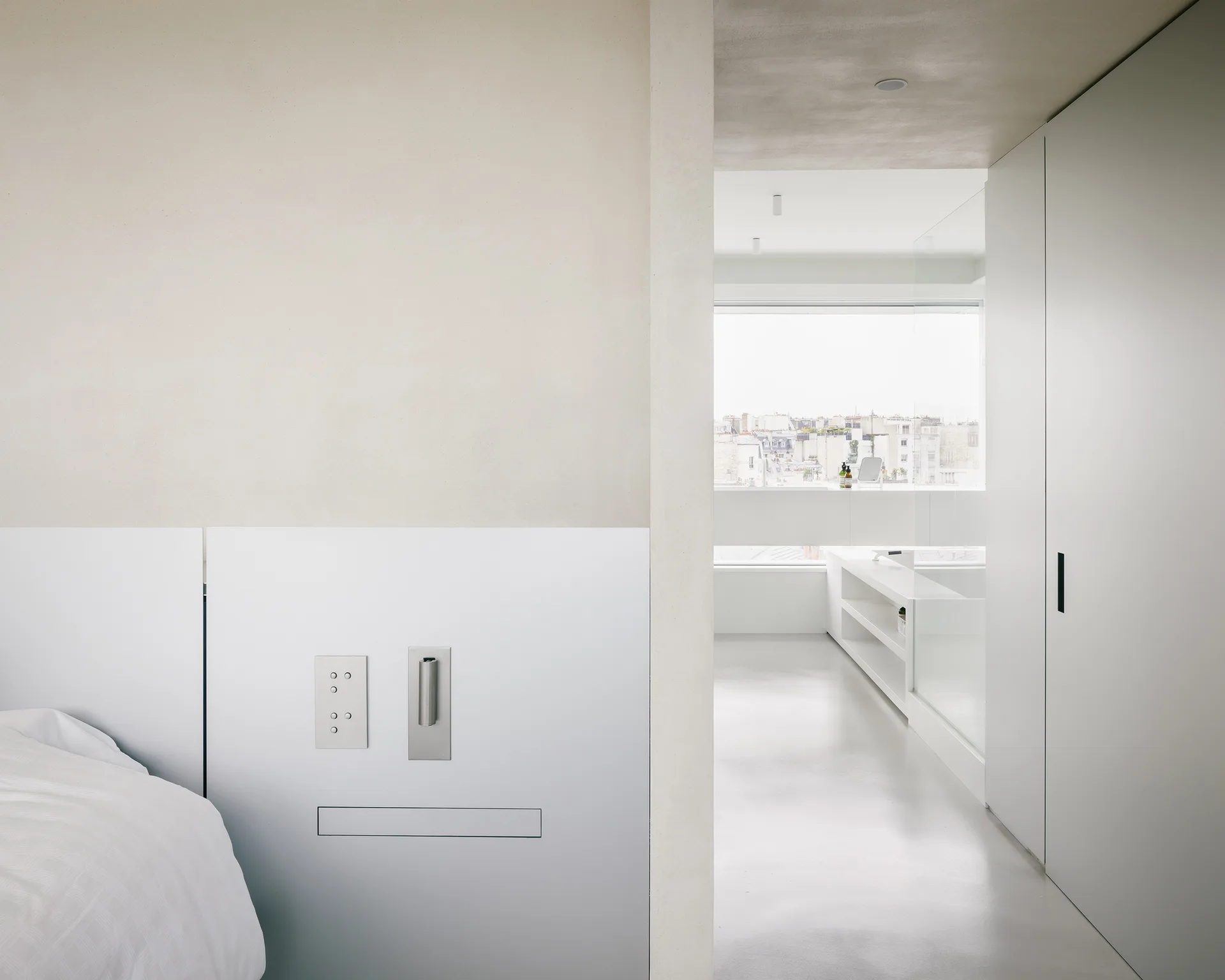 An off-white minimalist bedroom features a smooth plaster wall with flush metal switches next to an open doorway leading to a bright bathroom.