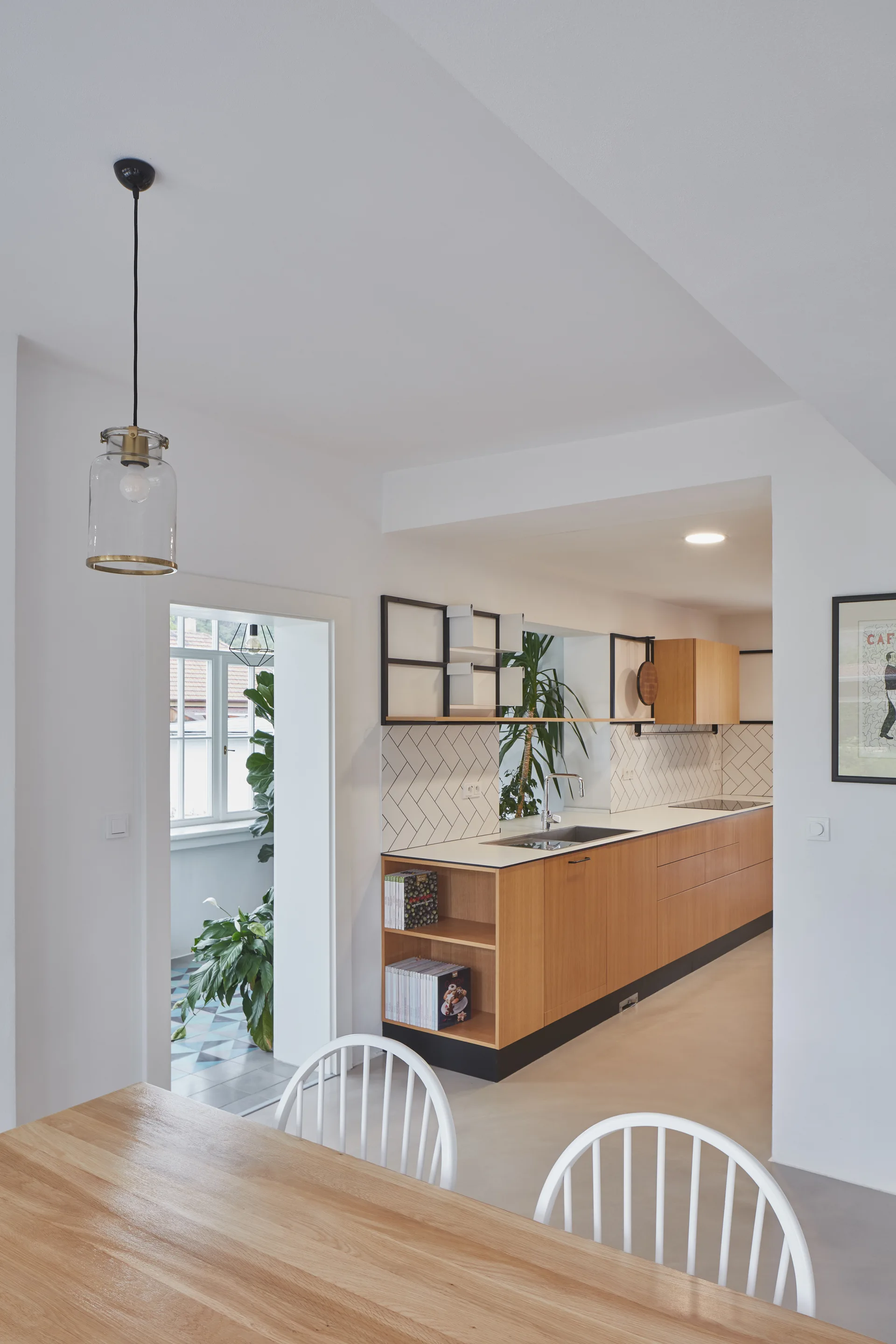 An open-concept kitchen features wood-toned cabinets, a white chevron tile backsplash, black open shelves, and a wooden dining table in the foreground.