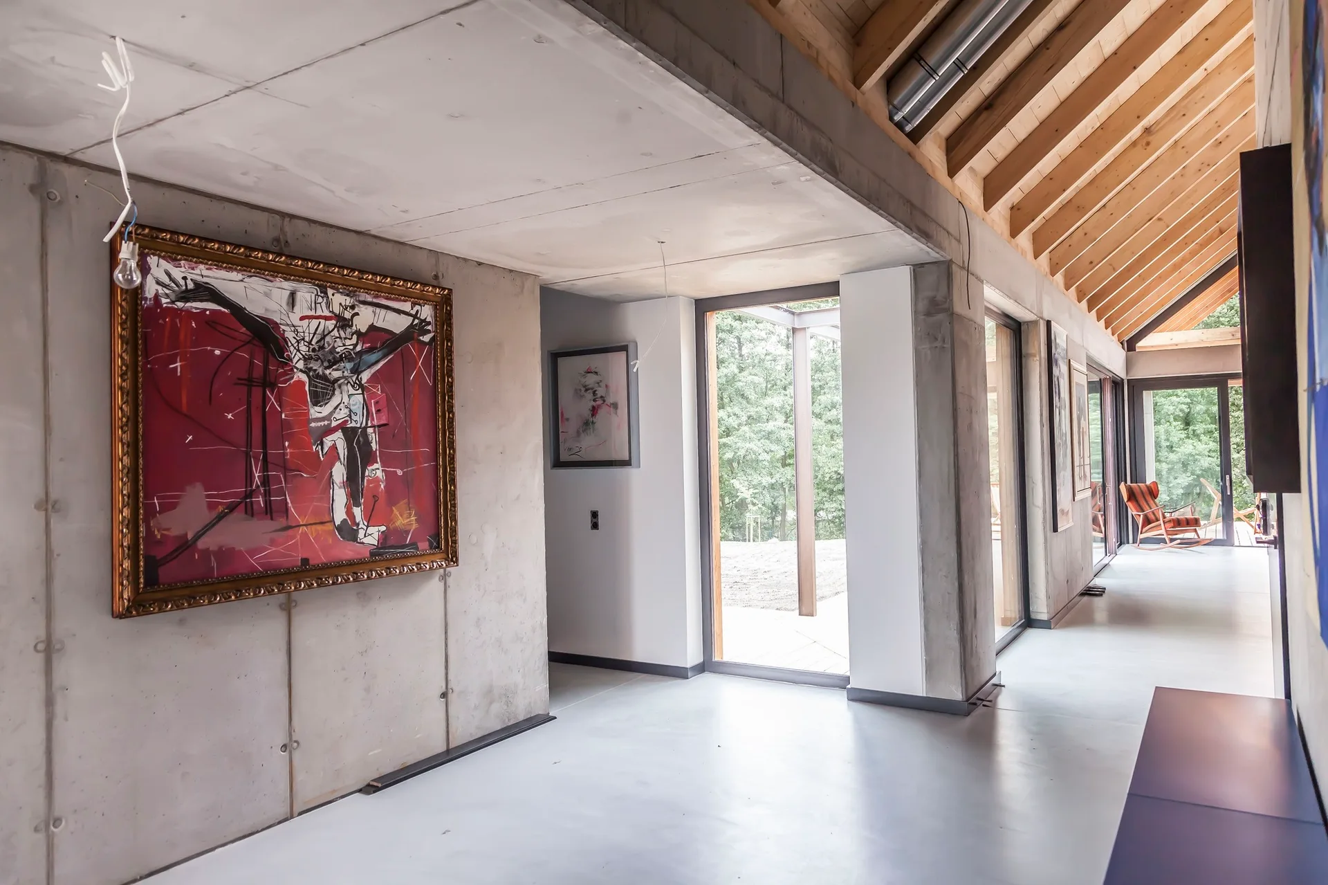 A minimalist hallway with raw concrete walls, exposed wooden ceiling rafters, floor-to-ceiling windows, and a large abstract red painting in a gold frame.