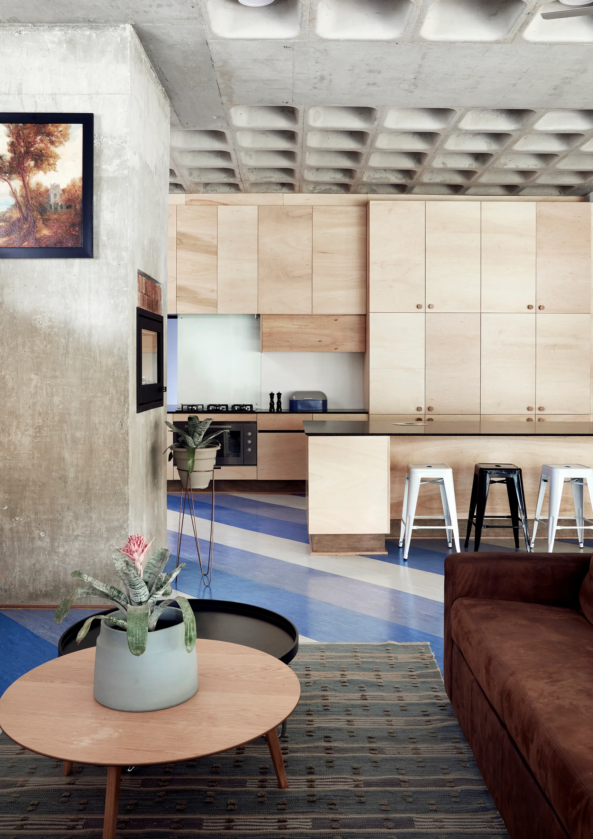 A modern open kitchen features light plywood cabinetry, an island with metal stools, an exposed coffered concrete ceiling, and blue striped flooring.