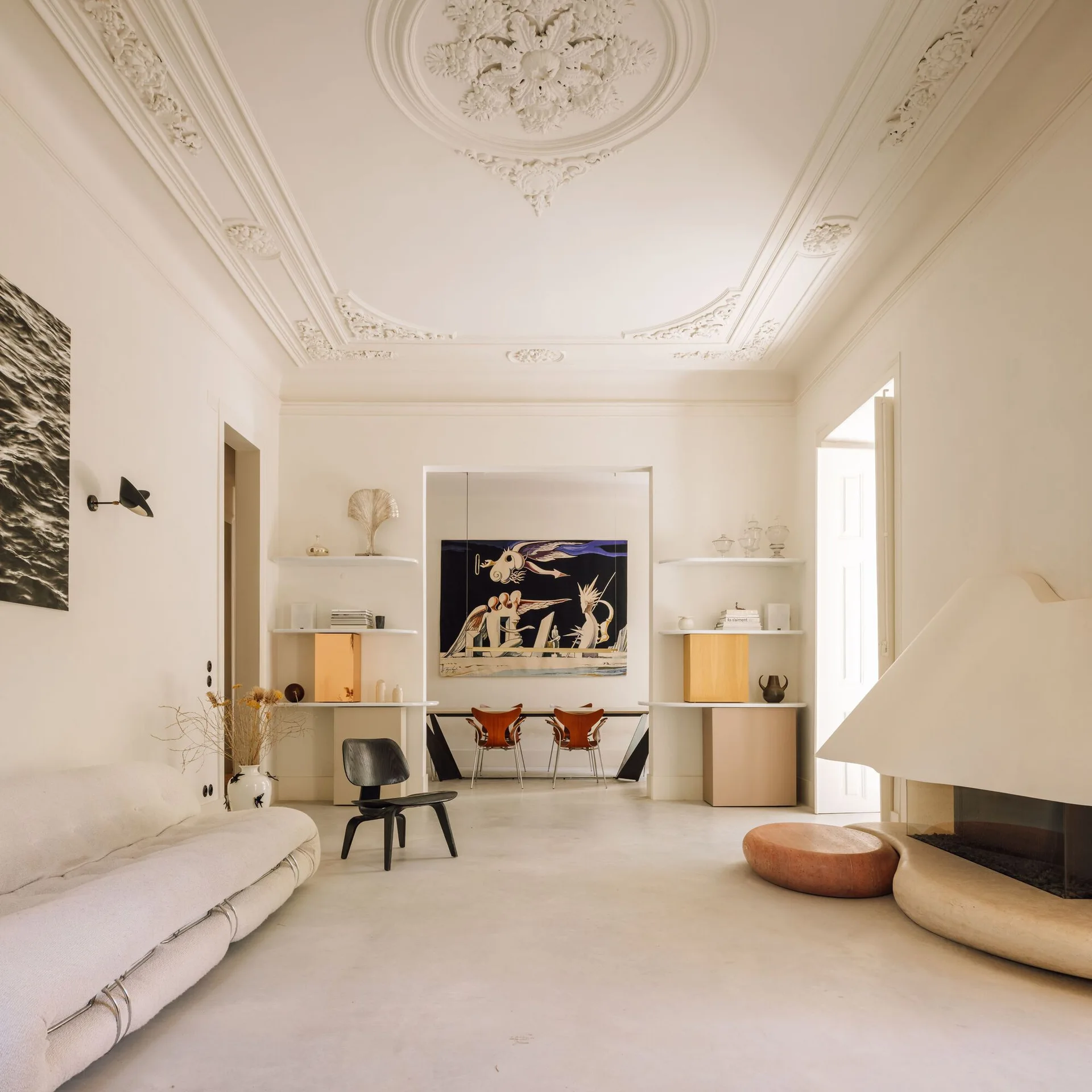 A bright living room featuring a sculptural plaster fireplace, low cream sofa, and built-in shelving beneath an ornate plaster ceiling.