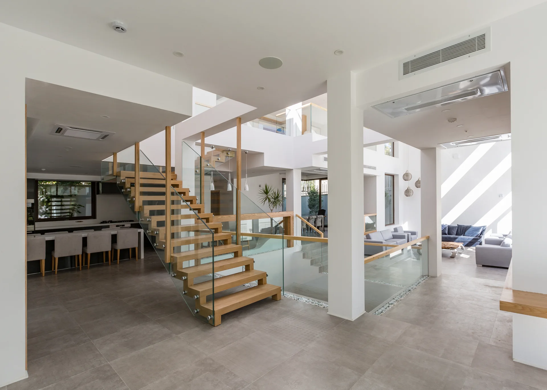 A spacious open-plan home interior features a central wooden staircase with glass railings, grey tiled floors, and white structural columns.