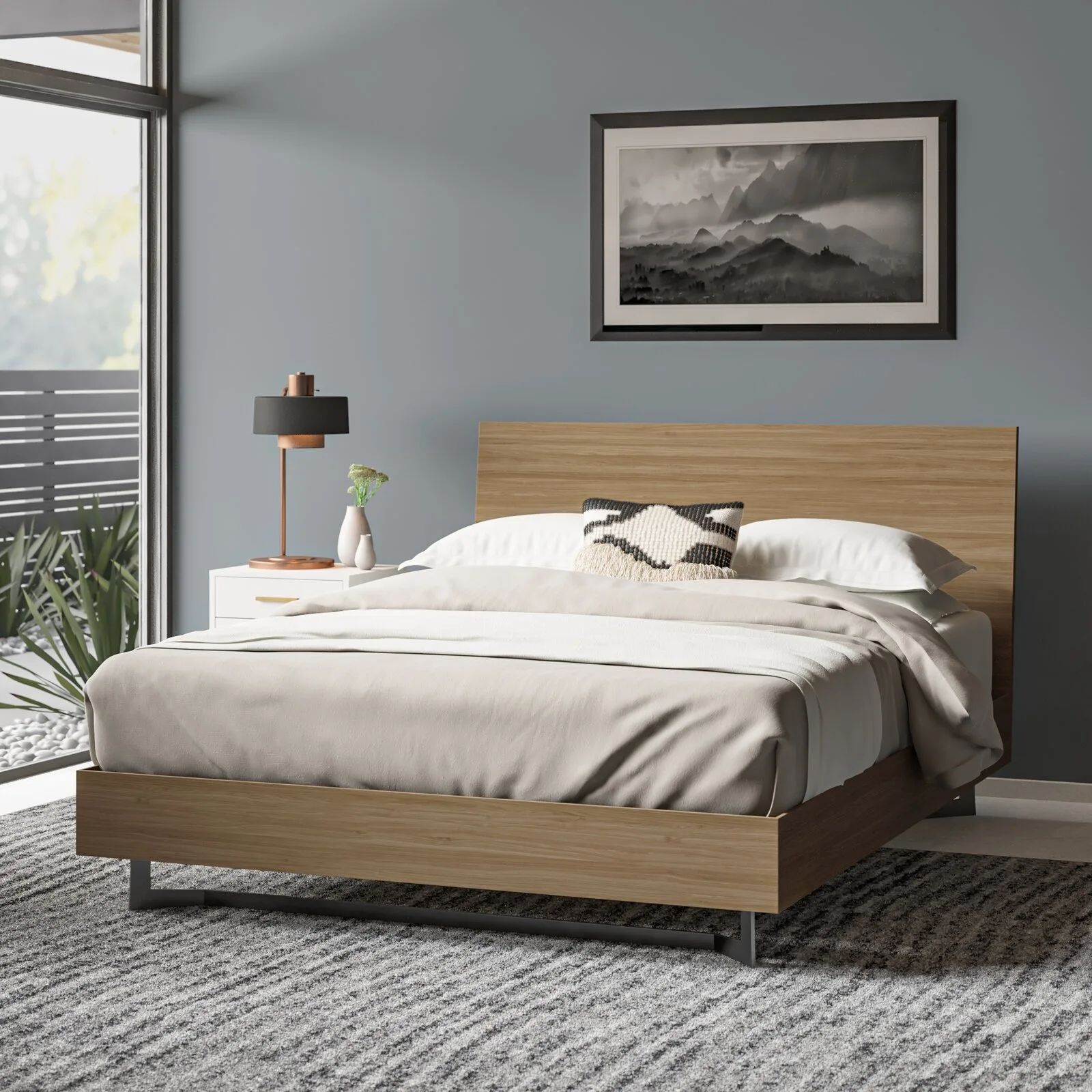 A modern bedroom features a warm wood platform bed on a textured grey rug, set against a muted blue-grey wall with framed landscape art.