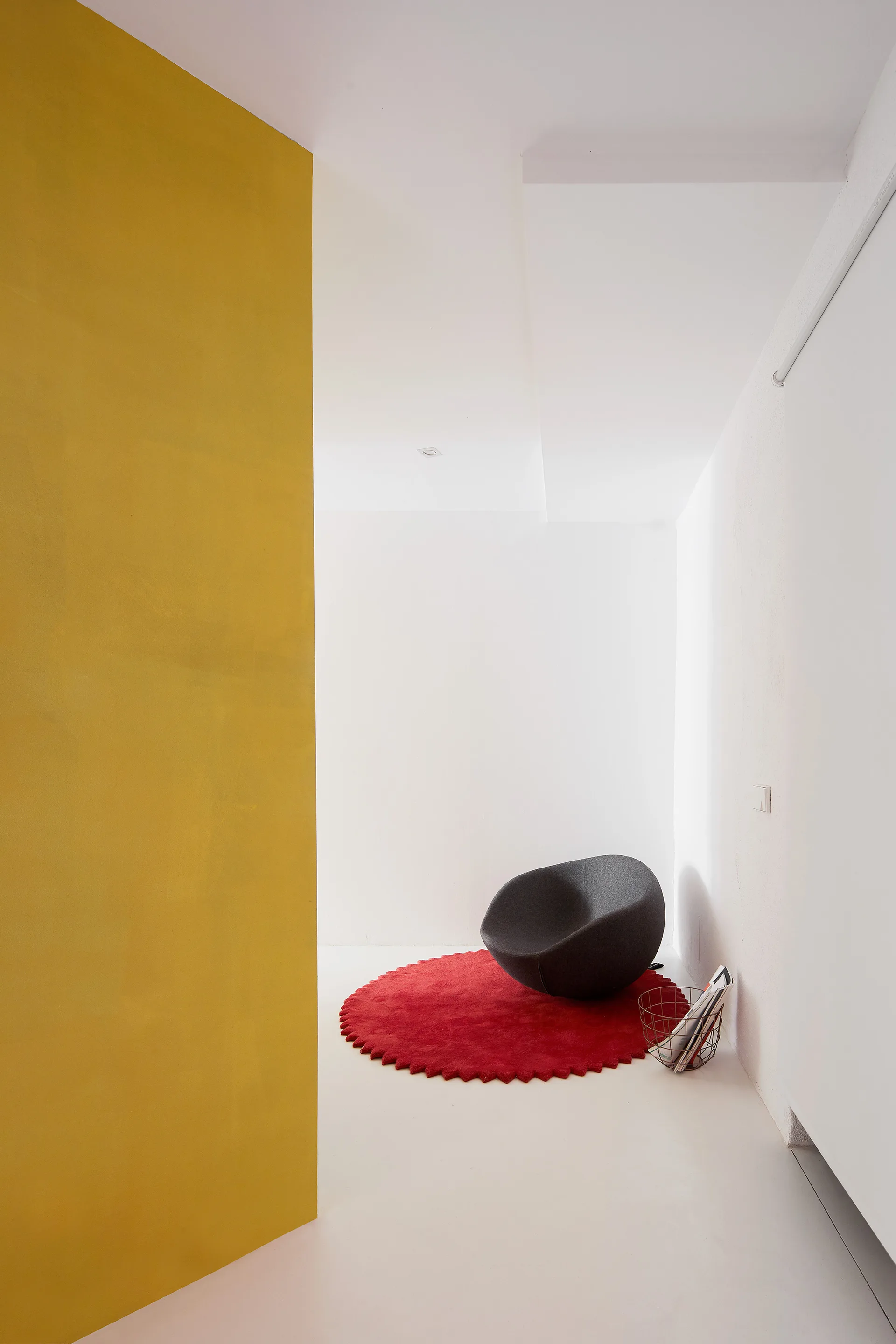A minimalist white room features a dark grey sculptural lounge chair on a red circular rug, framed by a prominent yellow wall segment.